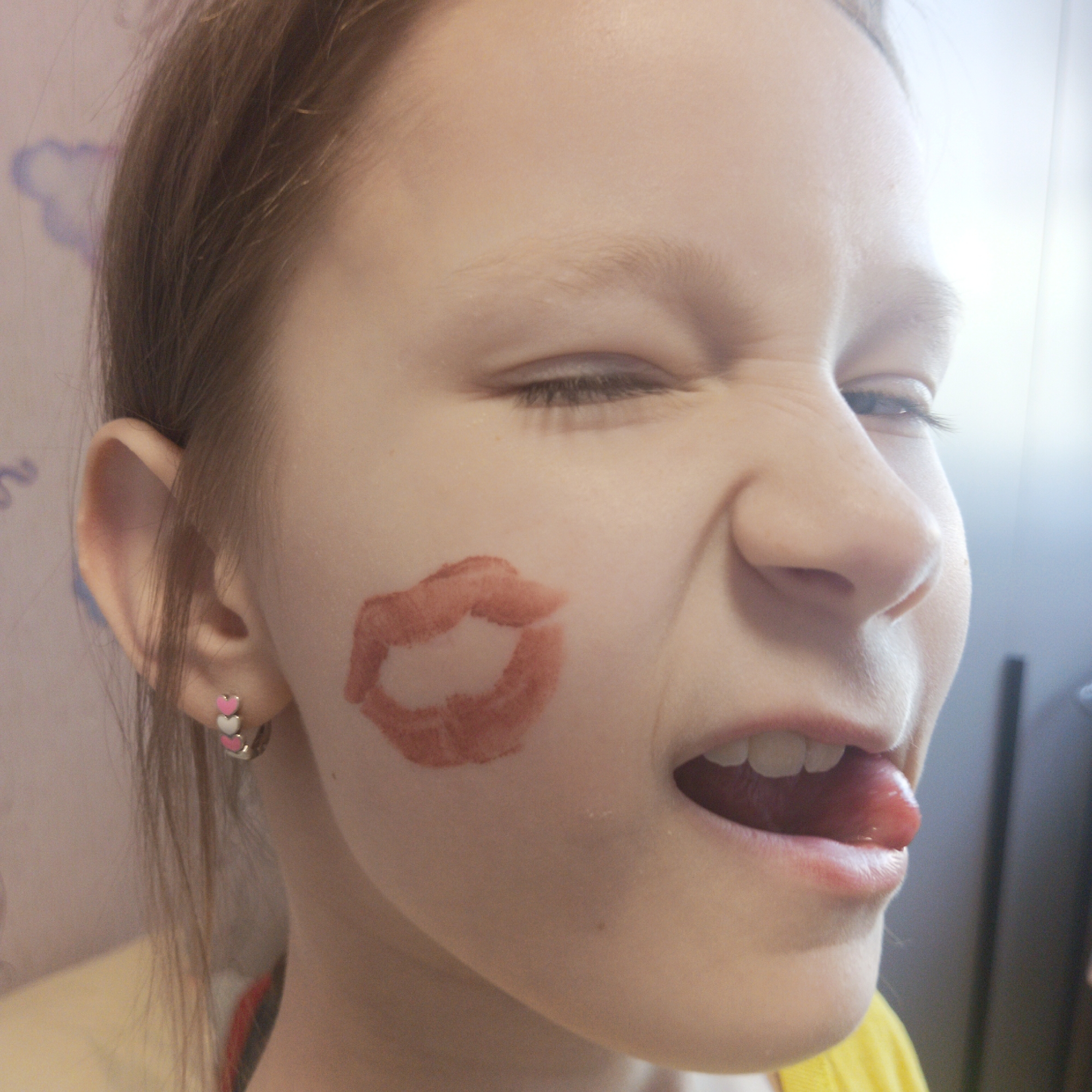 A young girl with fair skin and light brown hair is depicted in a close-up shot. She is making a face, squinting one eye and sticking out her tongue. A lipstick mark in the shape of lips is visible on her right cheek. She is wearing hoop earrings adorned with small pink and white hearts. The background appears to be an indoor setting, possibly a room with light-colored walls. A cloud-shaped design is faintly visible on the wall. While the image has the label "Chaikovsky, Russia", there is nothing in the image itself that explicitly confirms that location.