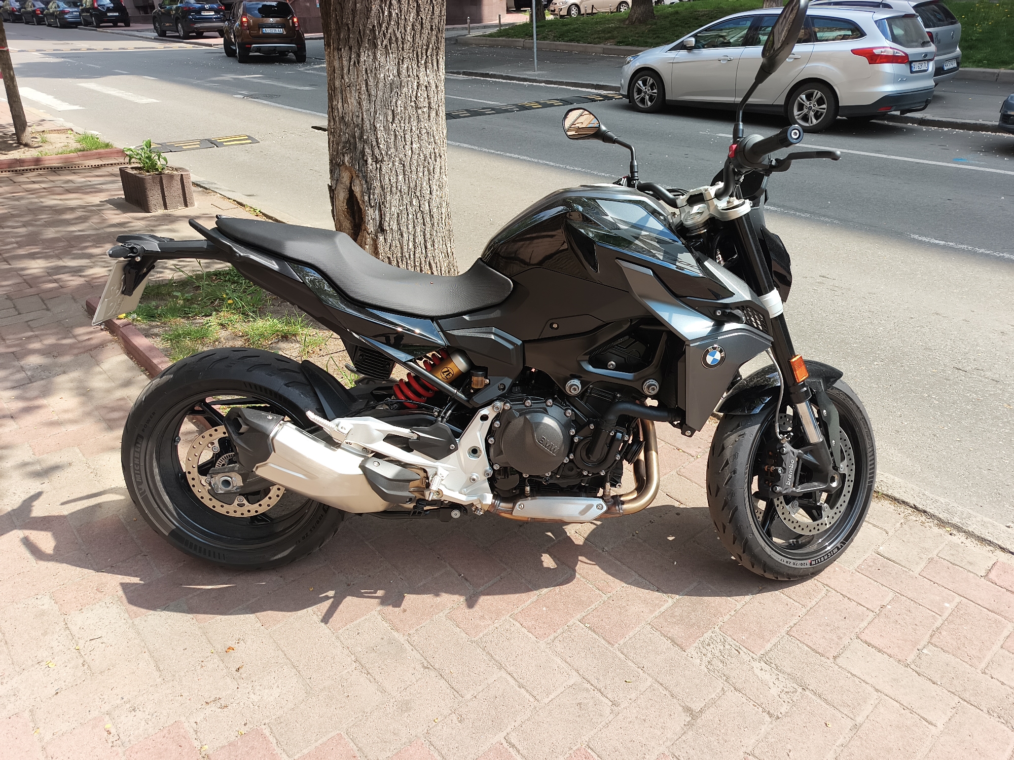 The image shows a black BMW F 900 R motorcycle parked on a brick sidewalk next to a tree, facing the right side of the frame. The bike is in focus, showcasing its design details such as the BMW emblem, Brembo brakes, and Michelin tires. In the background, a street with parked cars and crosswalk markings is visible, indicating an urban environment. Based on the license plates of the vehicles in the background, the location is in Kyiv, Ukraine. The weather appears to be sunny, evidenced by the shadows cast by the motorcycle and the tree. No people are visible in the frame.