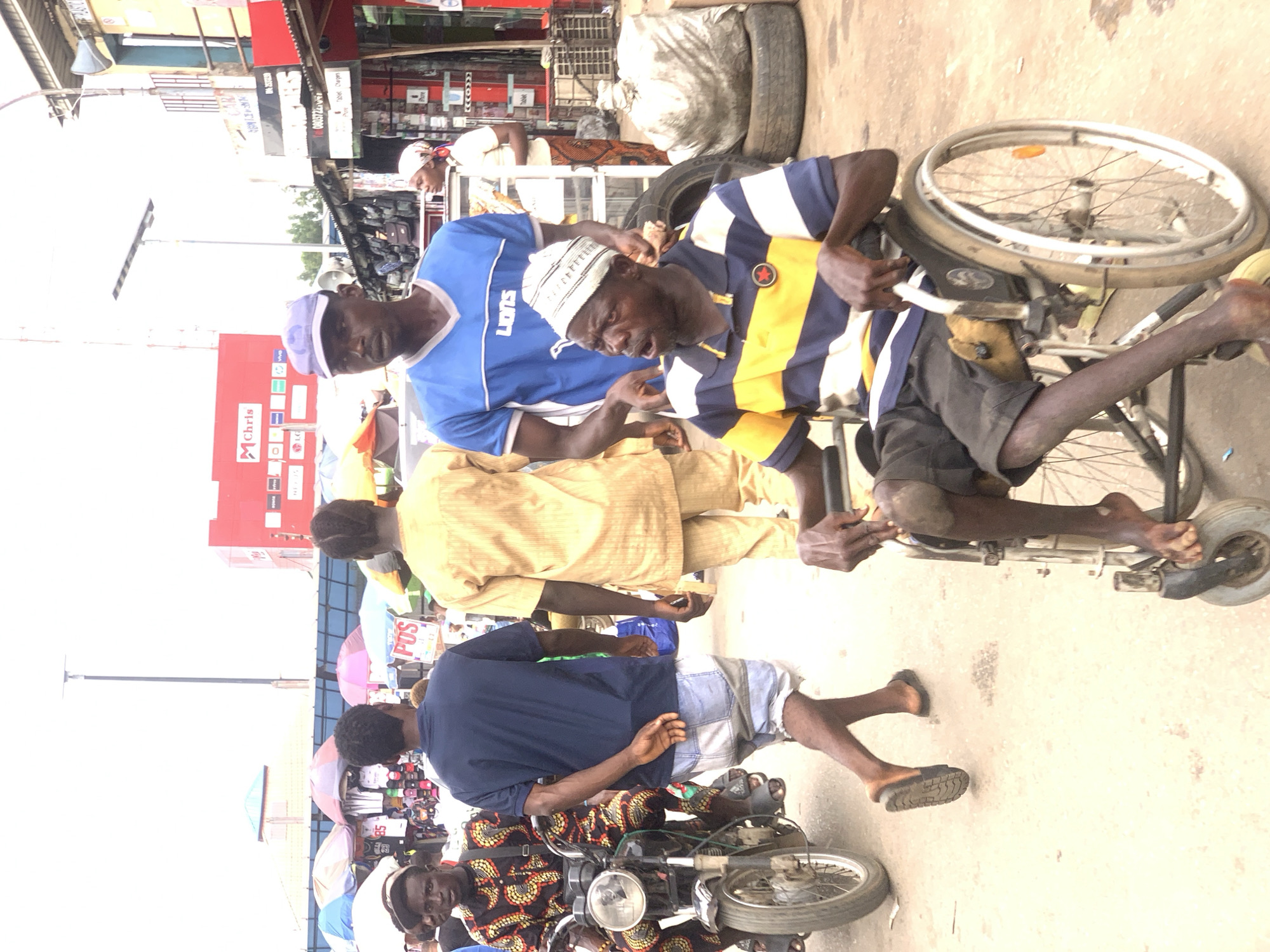 A man in a wheelchair is assisted by another man, possibly through a crowded outdoor market area. The wheelchair-bound man wears a blue and yellow striped shirt and a white cap. The man assisting him wears a blue t-shirt and a purple cap. Several other people are nearby, including a man in a patterned shirt on a motorcycle. Stalls with umbrellas are visible, suggesting a market setting. In the background, store signs read "Chris" and "POS". The image depicts a scene in Ikorodu, Nigeria, likely a bustling commercial area.