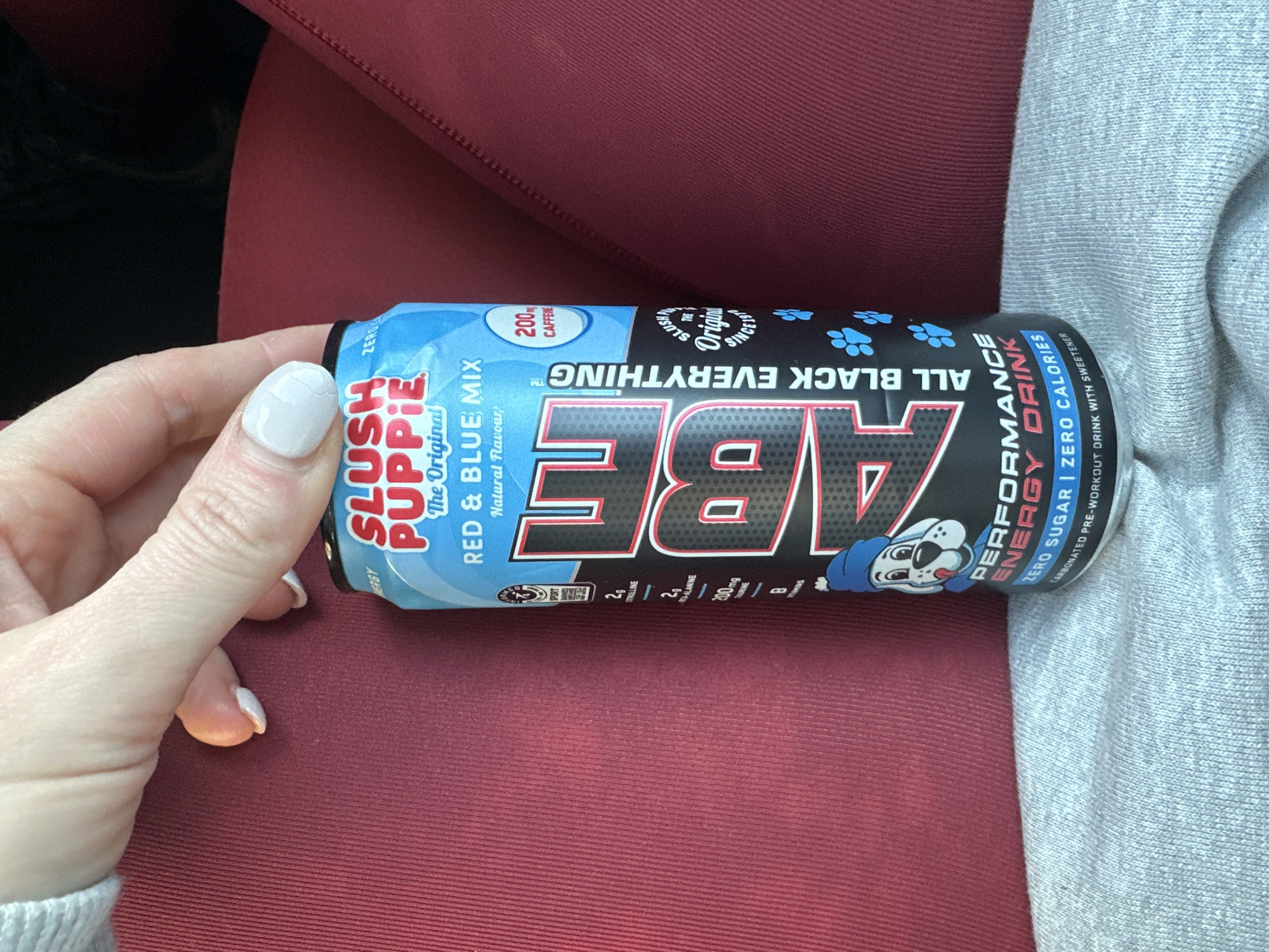 A light-skinned hand with white manicured fingernails holds the top portion of a can of ABE (All Black Everything) Performance Energy Drink. The can is labeled "SLUSH PUPPIE The Original Red & Blue Mix" and indicates "200mg CAFFEINE," "ZERO SUGAR," and "ZERO CALORIES." A cartoon dog mascot wearing a blue hat is depicted on the lower front. The can rests against a dark red, ribbed fabric surface, likely upholstery, with a lighter grey knitted fabric visible beneath it on the left. The background is indistinct, and the specific location within the city of Wealden, United Kingdom, cannot be determined from this image.