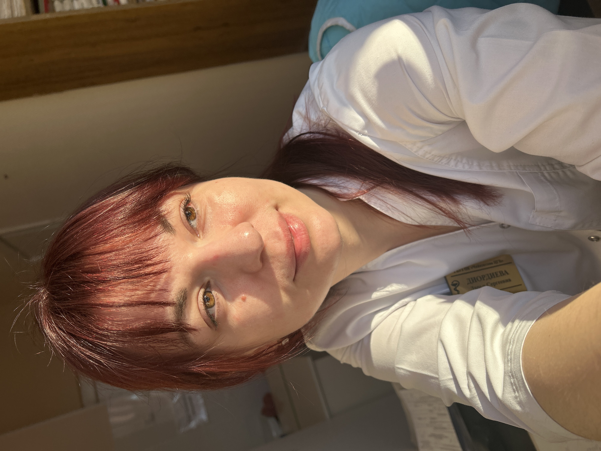 A woman with reddish-brown hair and light skin is wearing a white lab coat and smiling. A badge on her coat reads "ДЮРДИЕВА Лариса Сергеевна." The location is Фоки, Russia.