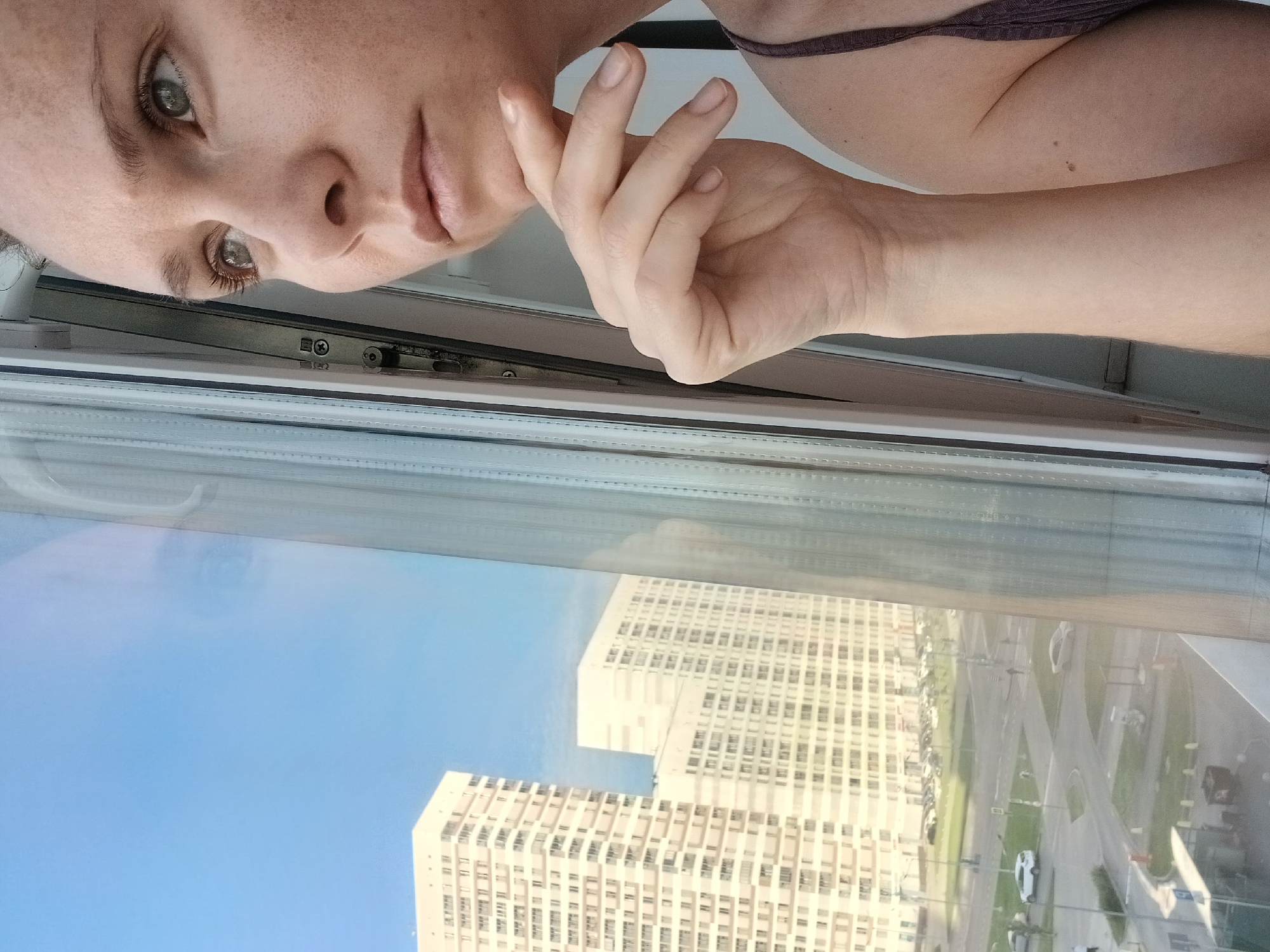 The image captures a woman looking out a window with buildings and a street visible in the background. The woman is in the foreground, her face partially obscured as she gazes to the left. Her right hand rests on her chin, and her arm is bent at the elbow. She appears to be wearing a dark-colored tank top. Reflected in the window are elements of the interior, including the window frame and a blurred light source. Outside the window, a clear blue sky is visible above several modern, tall residential buildings. Below the buildings, a street is discernible with parked cars and a few pedestrians. The overall scene suggests an urban setting, possibly a residential area with high-rise apartments.