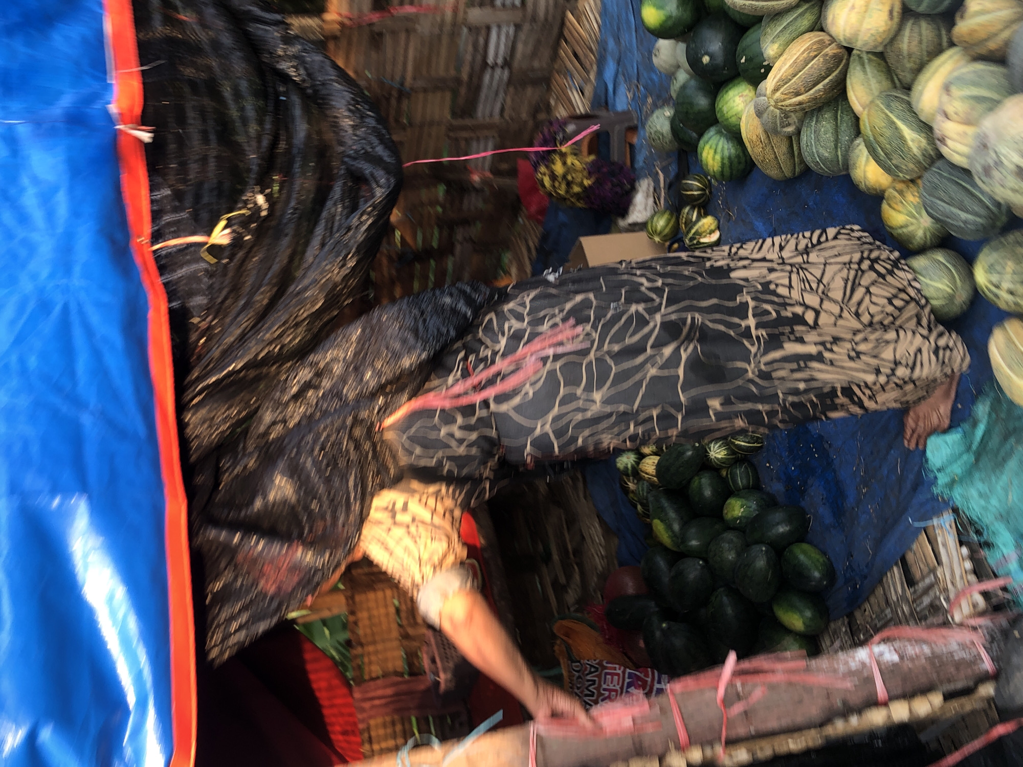 A street vendor in Glagah Arum, Indonesia, displays a variety of fruits, primarily watermelons and melons, on a blue tarp. A person, partially obscured by a black garment with a cream-colored abstract pattern, is visible behind the fruit display. The vendor's arm is extended, reaching towards the fruits. The scene is covered by a blue tarp, with red trim visible at the top edge. The backdrop appears to be a stall made of bamboo or similar natural materials. The overall image is slightly blurred due to motion.