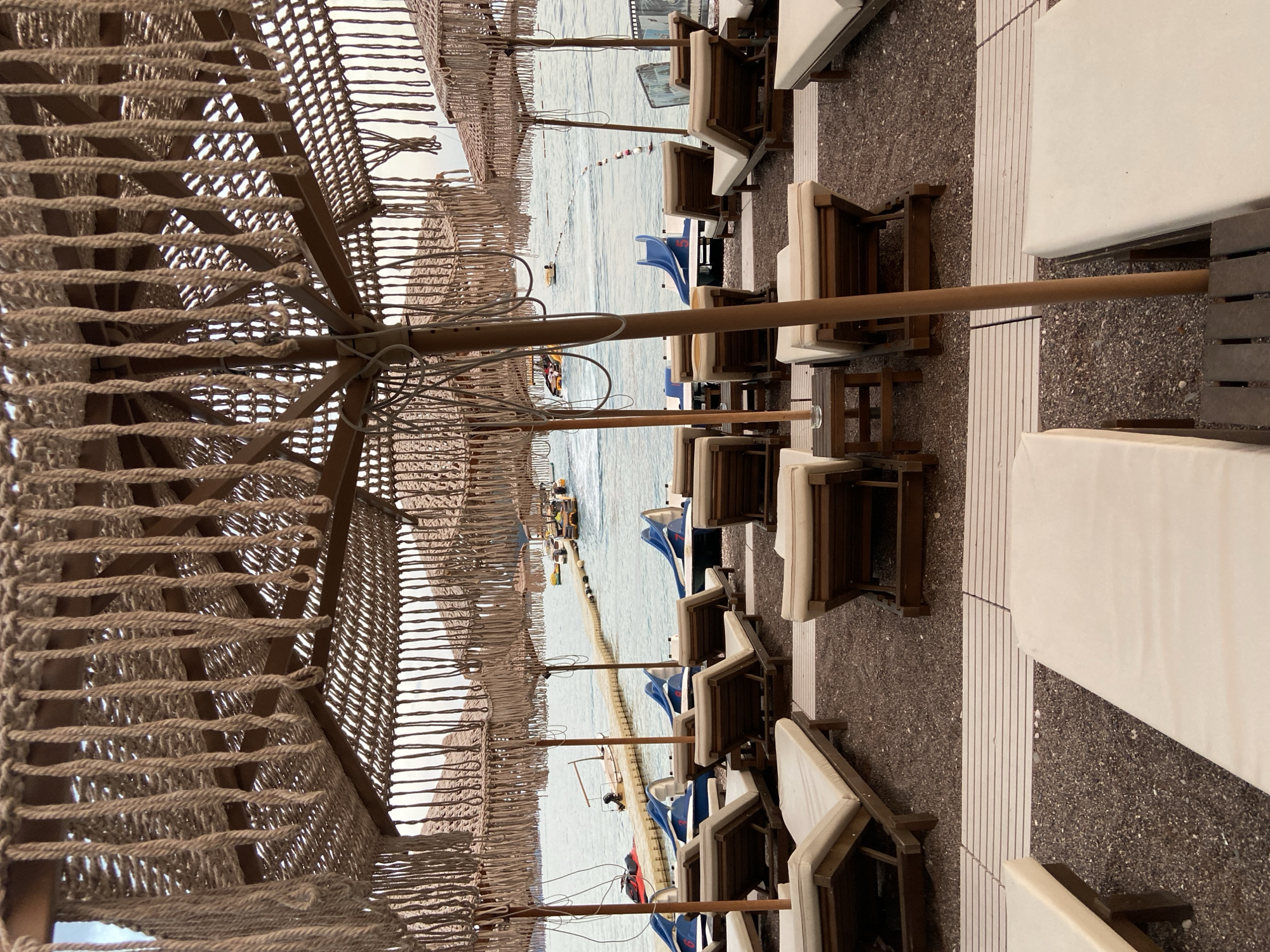 The image depicts a beach scene in Budva, Montenegro, characterized by rows of unoccupied lounge chairs arranged on a pebbled shore. Several beach umbrellas with woven rope detailing provide shade. In the background, the calm sea is visible, featuring a floating walkway with several paddle boats and a jet ski. A small boat is also anchored near the walkway. The sky is overcast. The setting appears serene and preparatory for beachgoers.