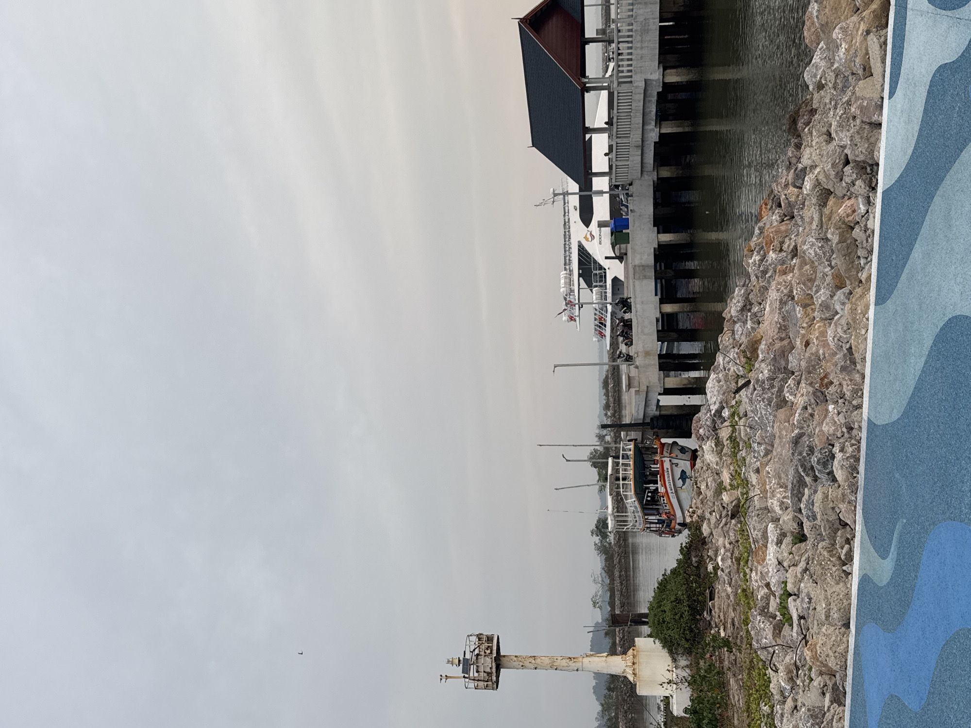 This image shows a port or harbor scene in Pak Nam Chumphon, Thailand. A small lighthouse stands at the foreground's base. Beyond this, two boats or ferries are docked. One, further away, is large and white. The other is smaller with an orange stripe. A building is connected to a pier where a boat is moored. Rocks are piled along the water's edge. Overcast sky conditions are evident. A blue and white patterned edge is visible on the right side, possibly part of a wall or walking path.