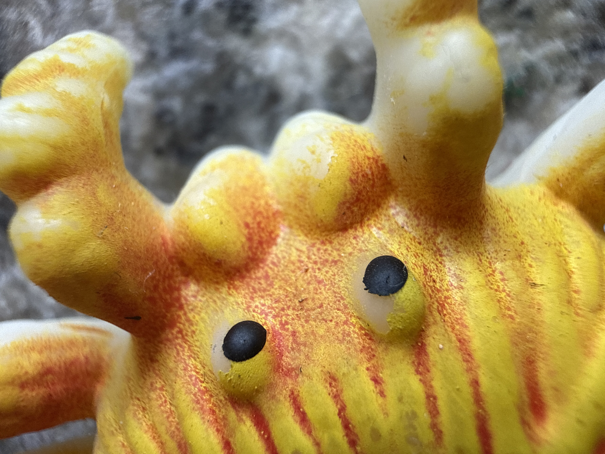 A close-up, macro photograph shows a yellow and orange plastic toy. The toy has two black, oval eyes embedded in its surface, which is textured and appears slightly fuzzy. Red stripes are painted across the yellow body of the toy. The background is out of focus and appears to be a rough, gray surface. There is no indication of the city of Ridabu, Norway, or any other specific location.