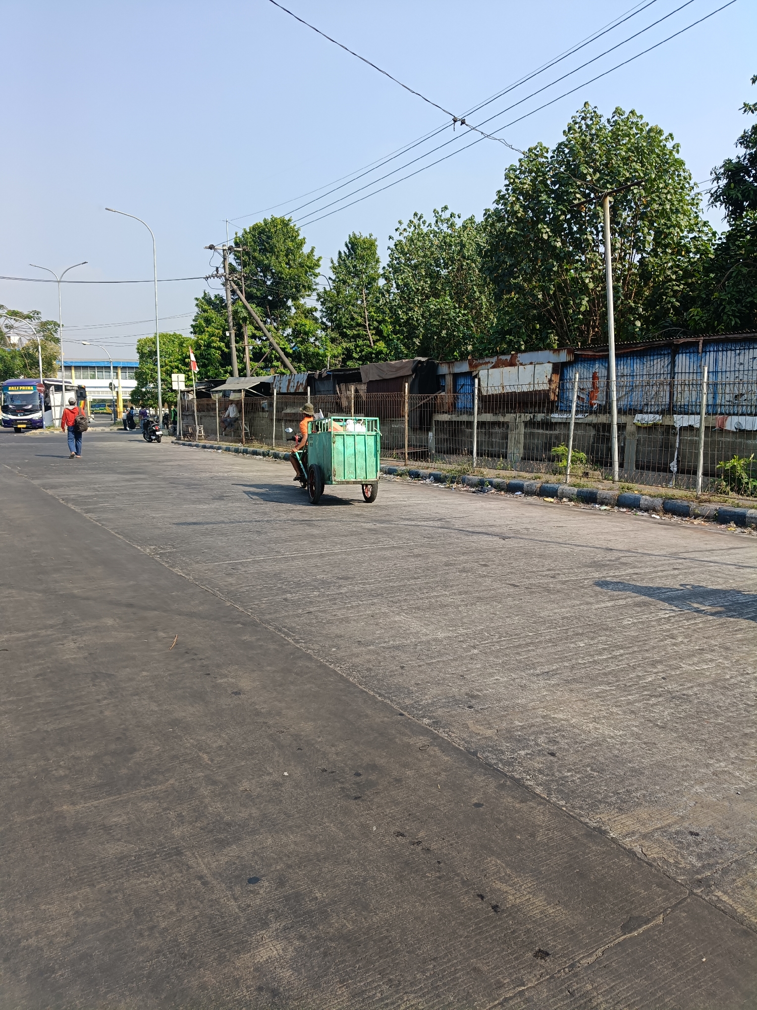 In the city of Kedungrejo, Indonesia, a man is riding a three-wheeled motorized vehicle with a green cargo box on a paved road. The vehicle is moving away from the camera, toward the right side of the frame. The man is wearing a hat and an orange shirt. The cargo box appears to contain various items, including white plastic bags.
The road surface is made of concrete and asphalt, with some debris scattered on it. To the right of the road is a fence constructed of metal poles and mesh, behind which are some structures with corrugated metal roofs and blue siding. Beyond these structures, a line of trees with dense green foliage is visible against a clear blue sky.
To the left of the road, further in the background, is a purple bus with "DALI PRIMA" written on its side. A person wearing a red hoodie and carrying a backpack is walking away from the camera on the left side of the road. Another person is riding a motorcycle in the distance on the left. Streetlights and power lines are also visible. The scene appears to be an urban or semi-urban environment with evidence of commercial or industrial activity.