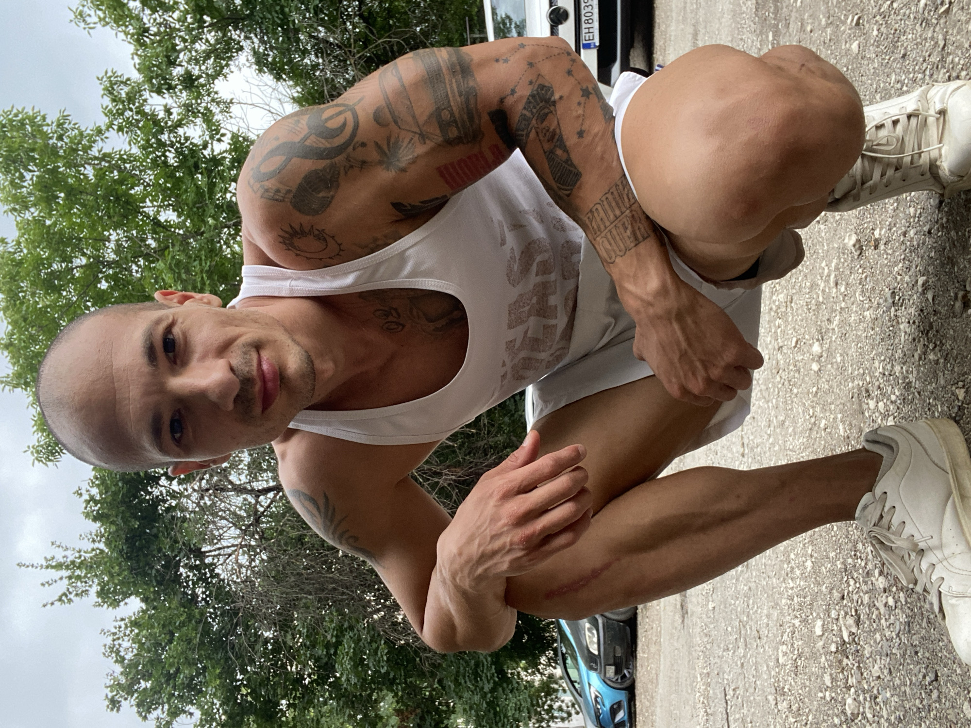A bald man with tattoos on his arms and leg kneels on a gravel surface. He is wearing a white tank top and white shorts. His left hand is resting on his left knee, which has a visible scar. In the background, trees and cars are visible. The scene is outdoors, under a cloudy sky. The location is identified as Pleven, Bulgaria, based on the license plate of one of the cars in the background.