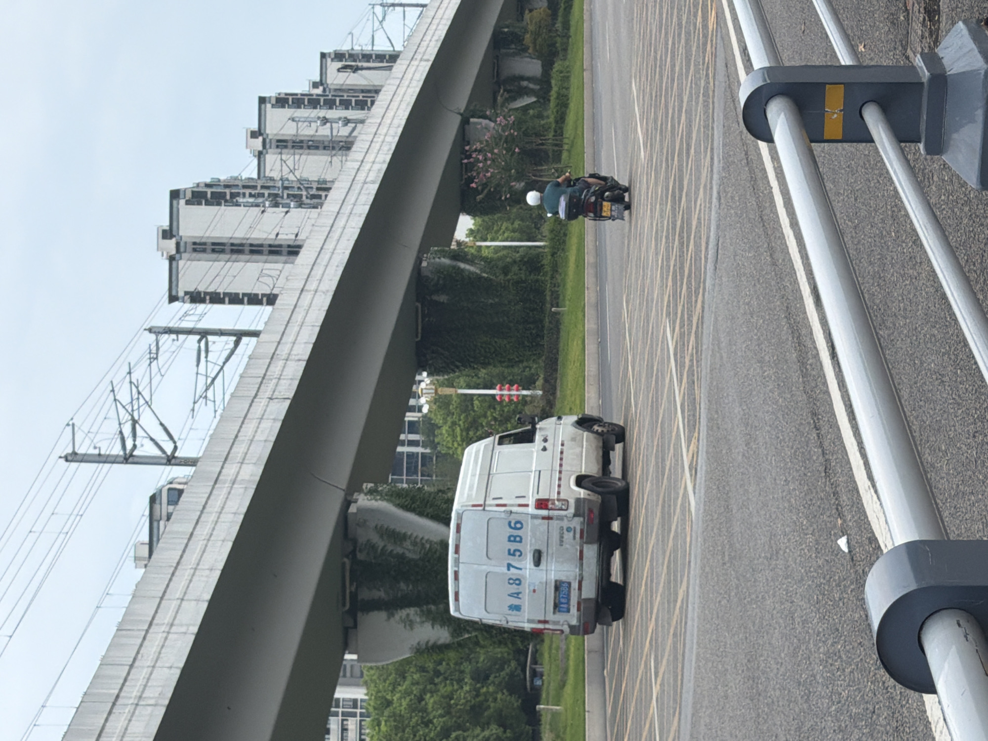 The image depicts an urban scene in Jiangjin District, Chongqing, China, with an elevated railway or highway structure dominating the background. In the foreground, a white van with the license plate "A875B6" is parked in a marked parking area with yellow and white lines. Further down the road, a person wearing a white helmet is riding a motorcycle. The environment includes green trees and residential buildings in the middle ground. The sky is overcast.