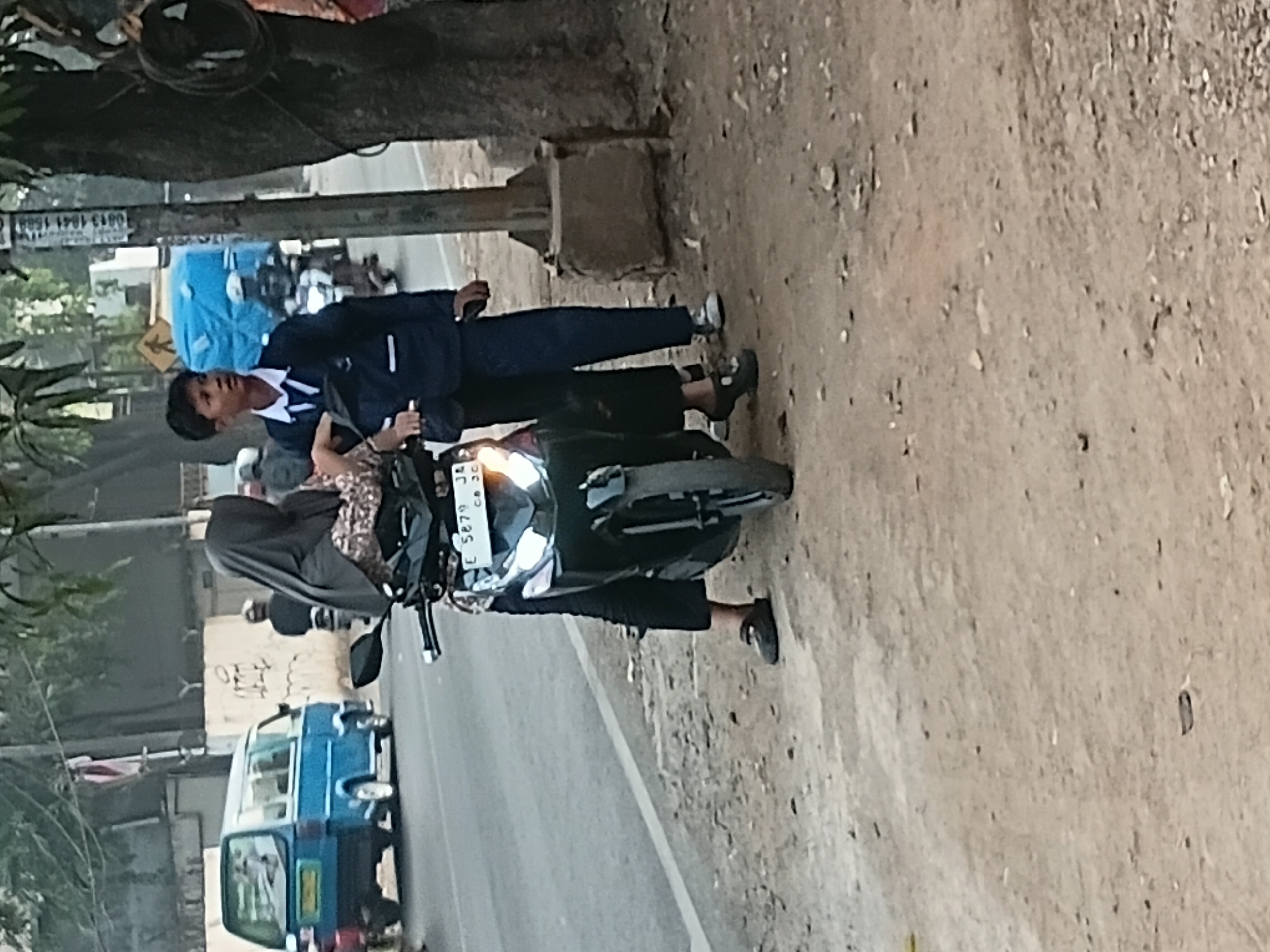 In the city of Bekasi, Indonesia, two individuals are standing next to a parked motorcycle on a dirt roadside. The person on the left, dressed in a black hijab and a patterned shirt, is seated on the motorcycle. The person on the right, a young male wearing a dark blue school uniform with a white collar and tie, stands beside the motorcycle, looking to the left. A blue minivan is visible in the background, driving on the street. A utility pole stands to the right of the individuals. The overall environment suggests a street scene with moderate urban activity.