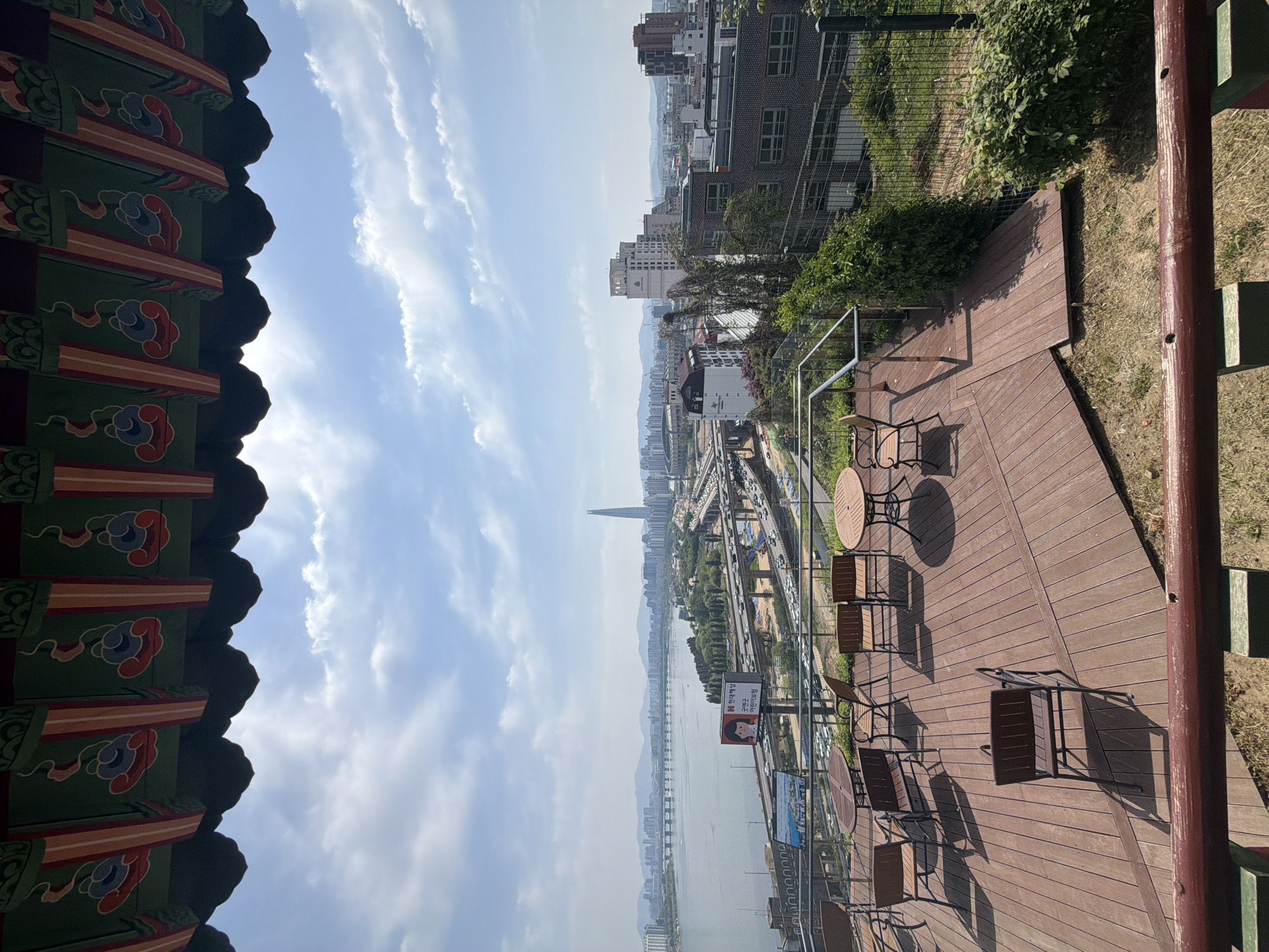 The image shows an elevated view in Seoul, South Korea, looking towards the Han River. The Lotte World Tower is visible on the horizon. The viewpoint includes a wooden deck with several small tables and chairs, suggestive of a cafe or outdoor seating area. A building with traditional Korean architectural details (a painted eaves element) is visible on the left side. The sky is partly cloudy, and the buildings of Seoul stretch out along the river. There are roadways with visible traffic. The deck is bounded by a low fence or barrier.