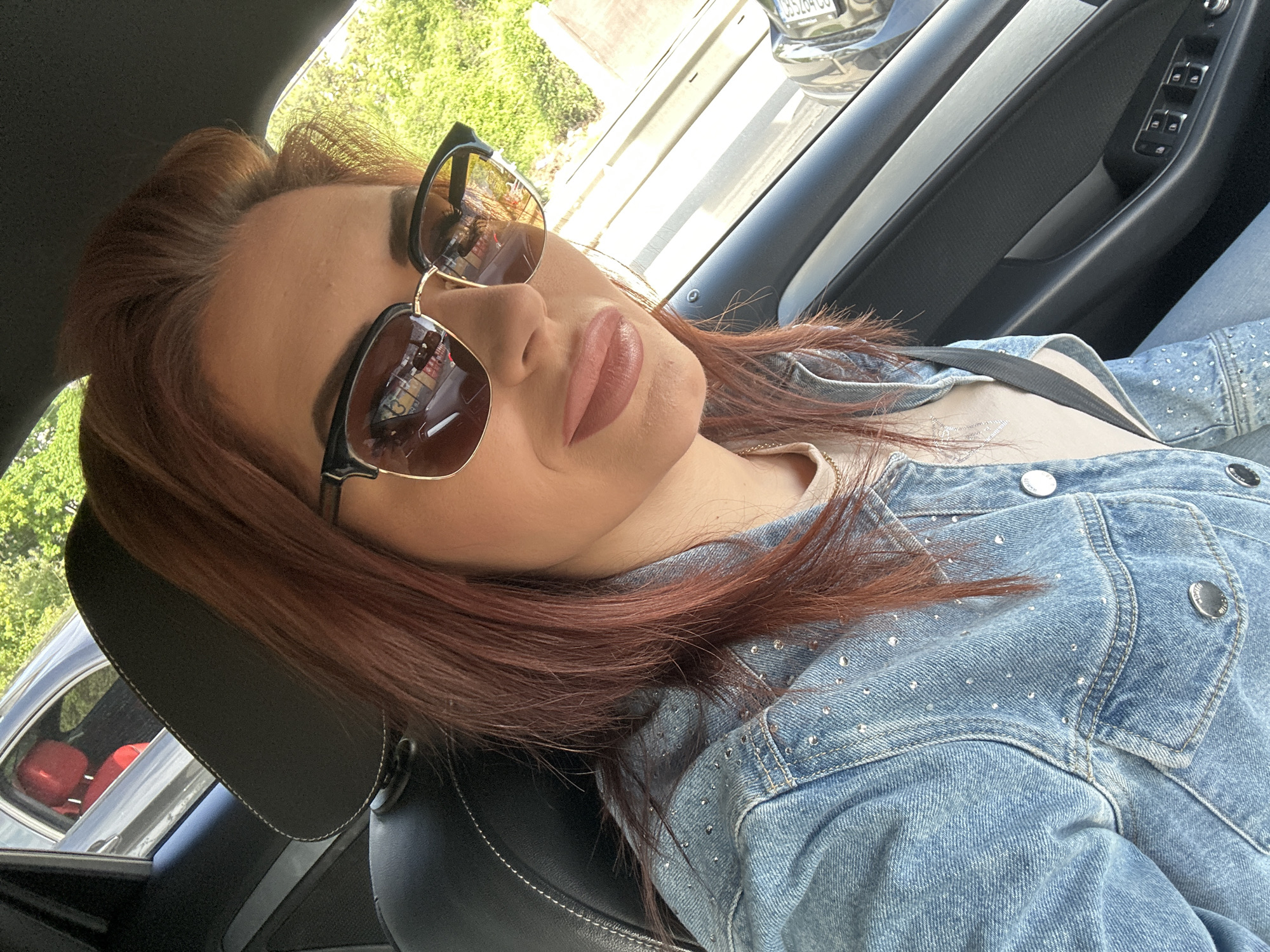A woman is in a car wearing sunglasses, a denim jacket with rhinestones, and a seatbelt. Based on the reflection in the sunglasses, the location is possibly Sofia, Bulgaria, as the license plate of a vehicle reflects a Bulgarian license plate format (e.g., "C RS 264 CC"). Trees and buildings are visible through the car window. The car's interior features black and silver trim.