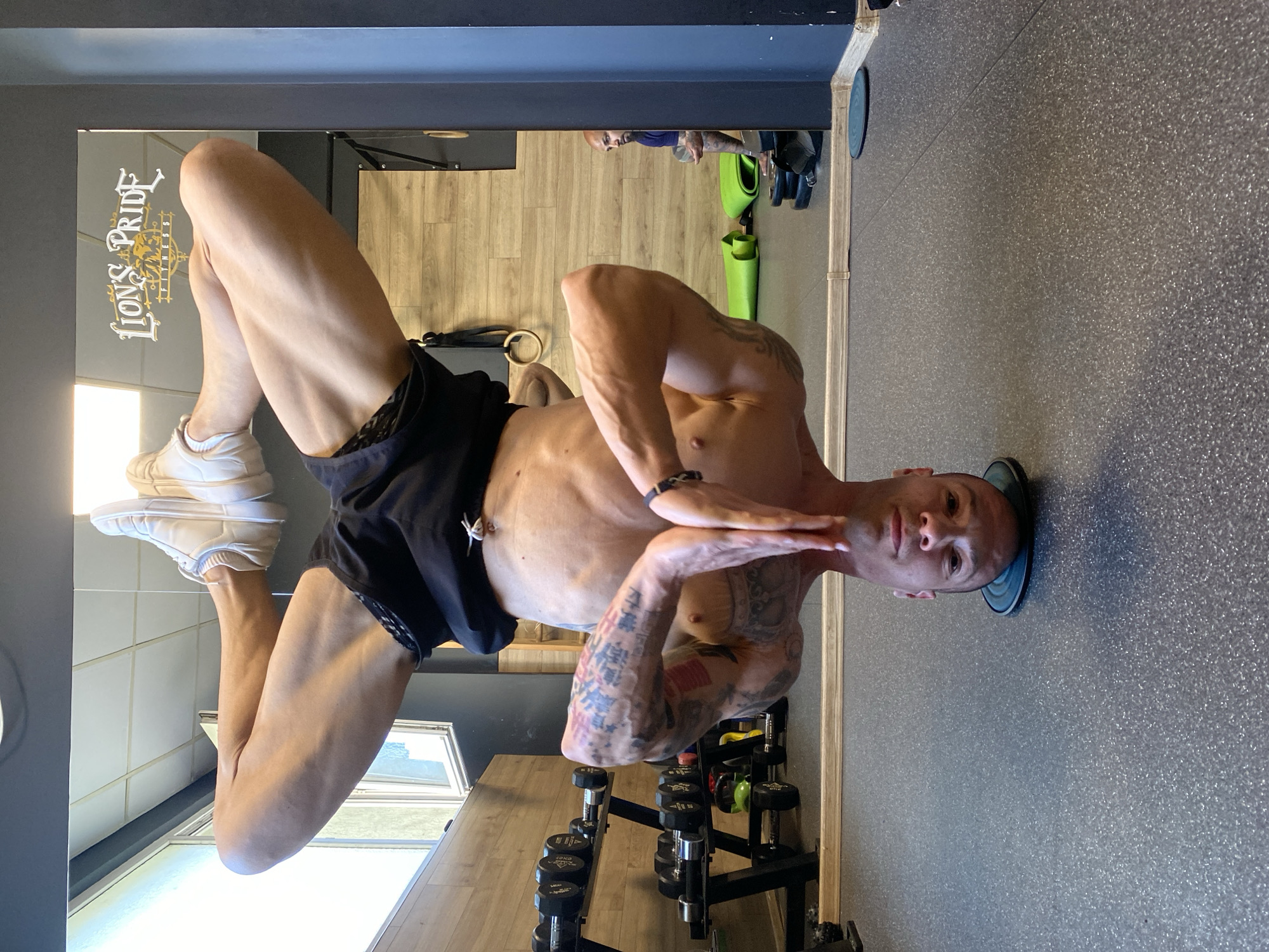 An athletic man is captured mid-exercise, performing an inverted bodyweight pose, possibly a headstand or a similar balance maneuver, within a gym in Pleven, Bulgaria. He is shirtless, showcasing a lean, muscular physique with prominent tattoos on his arms and torso. He wears black athletic shorts and white sneakers, with his head resting on a small, circular black and blue object on a speckled grey floor. His hands are positioned in front of his chest, and his expression is one of focus and concentration.
The background reveals typical gym equipment and features. A large mirror reflects sections of the gym, including a light-colored wooden floor, several green rolled-up yoga mats, and a dark wall or beam. A black rack of dumbbells is visible, with one clearly marked "10KG". The reflection also shows part of another person in the distance. The gym's name, "LIONS PRIDE FITNESS," is prominently displayed on the mirror in a decorative, golden script. The lighting suggests it is daytime.