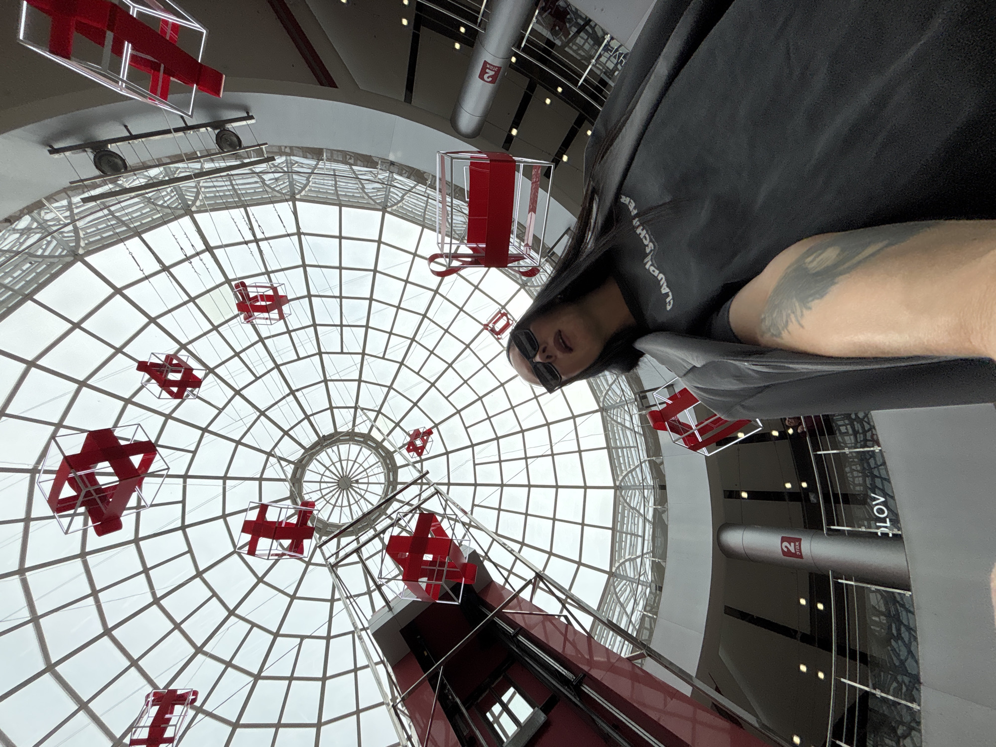 A tilted, upward-angle shot shows a person with dark hair and sunglasses in the lower-right portion of the frame, their arm visible. The backdrop is a circular, glass-paneled ceiling with red abstract airplane structures hanging within the space. A red elevator structure is also visible in the lower portion of the image. The scene is identified as the interior of the shopping center "TALISMAN" in Izhevsk, Russia. A cylindrical pillar with the number "2" on it can be seen.