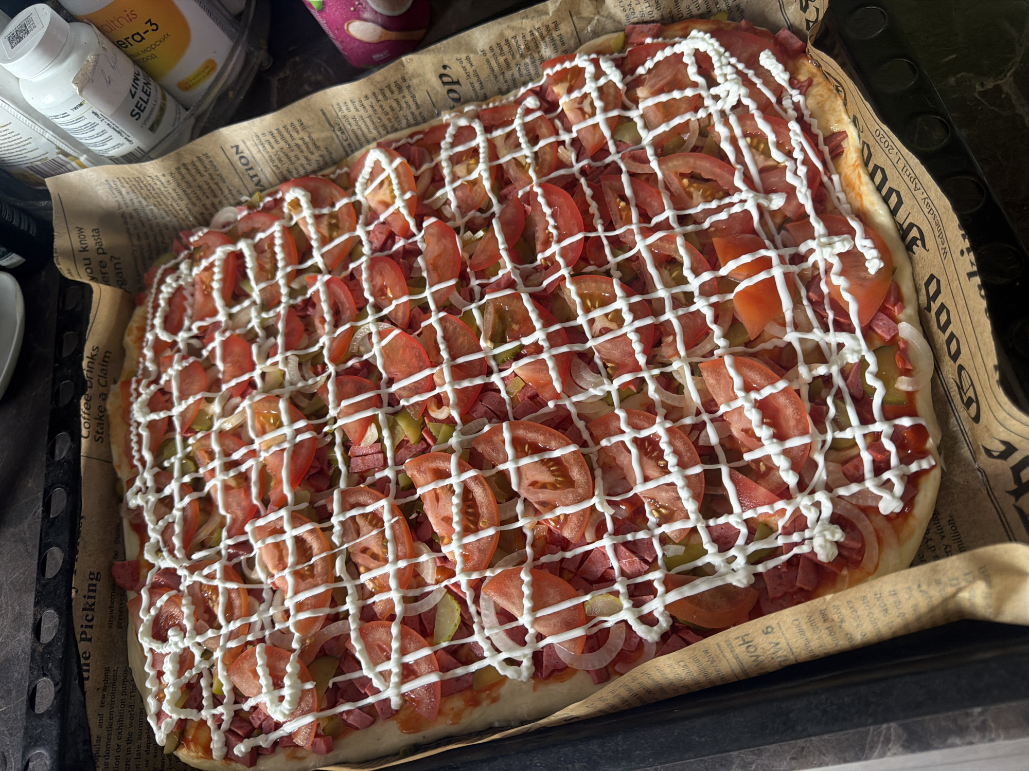 The image shows a rectangular pizza in a baking pan lined with newspaper. The pizza has a visible crust and is topped with sausage slices, tomato slices, onion rings, and pickles. A grid-like pattern of white sauce (possibly mayonnaise) covers the toppings. Next to the baking pan are containers of pills and other food items. The newspaper used as a lining is dated Wednesday, April 1, 2020 and has English text on it. The exact location is difficult to determine without additional details, but considering the Cyrillic text "Срок годности 2 года" on a container, it may be from Russia.