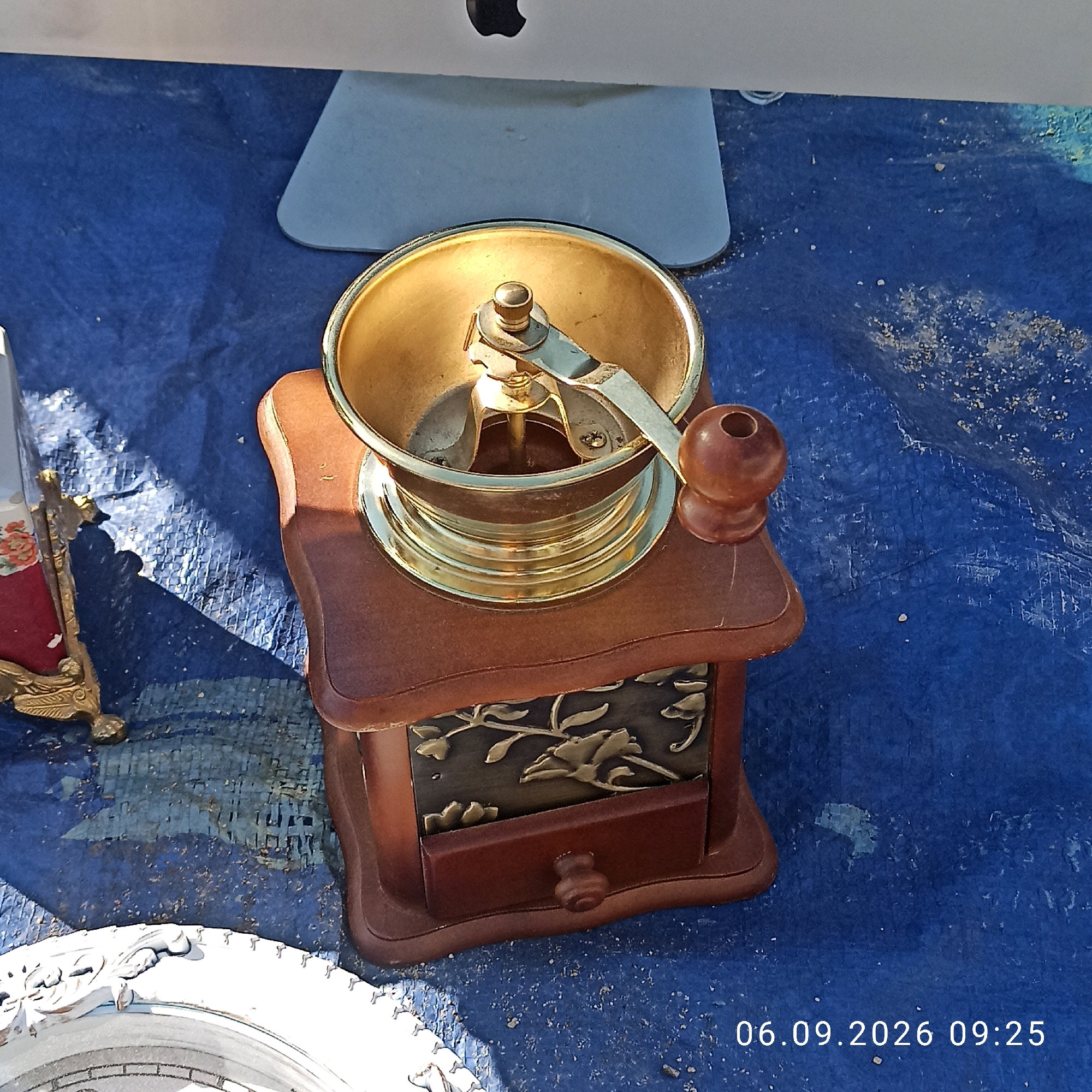 A wooden and brass manual coffee grinder sits on a blue tarp. The grinder has a wooden base with a decorative metal inlay and a small drawer. The top part of the grinder is brass, with a crank handle on the side. In the background, a portion of an Apple computer monitor is visible. The scene appears to be outdoors, with some scattered debris on the tarp. The date and time stamp "06.09.2026 09:25" is visible in the lower right corner. The image's metadata indicates it was taken in Sannicandro di Bari, Italy.