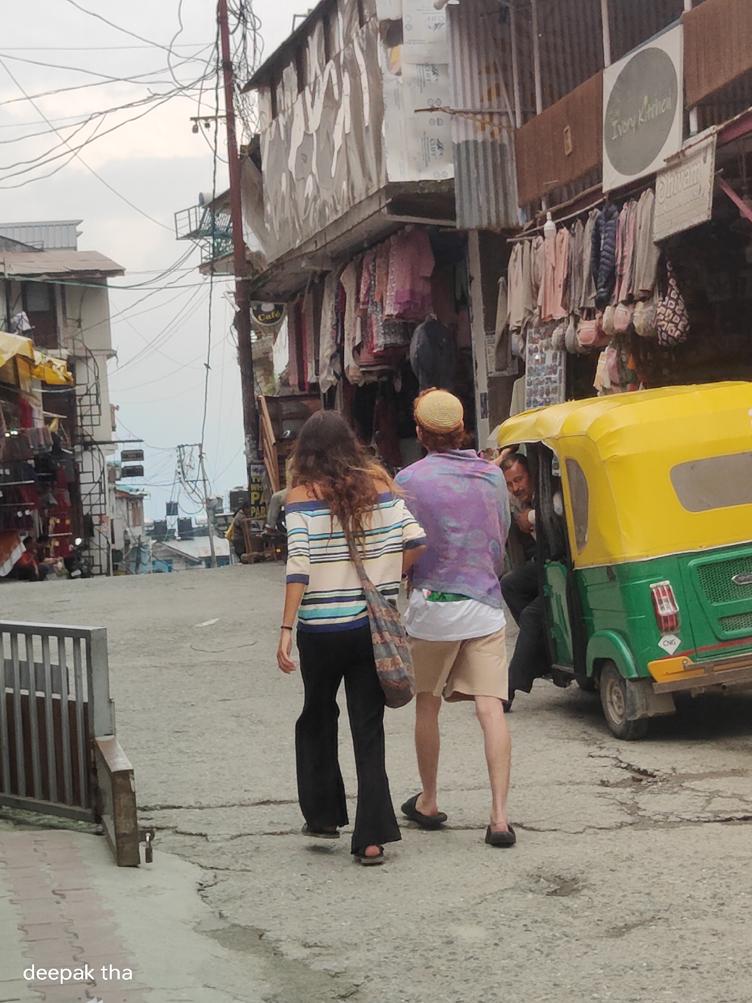 In Vashisht, India, a narrow, paved street slopes upwards between buildings on either side. Overhead, a dense network of electrical wires crisscrosses the sky. On the left, a building with corrugated metal siding displays an array of merchandise. On the right, shops with awnings showcase clothing and bags for sale. A yellow and green auto-rickshaw is parked on the street, and a man with a mustache is visible inside the driver's seat. A woman with long, wavy brown hair is walking away from the camera, wearing a striped long-sleeved shirt and black pants. Beside her, a person wearing a purple patterned shirt and tan shorts walks with them. The ground is textured with cracks and uneven patches. The overall atmosphere is that of a bustling marketplace in a hilly area.