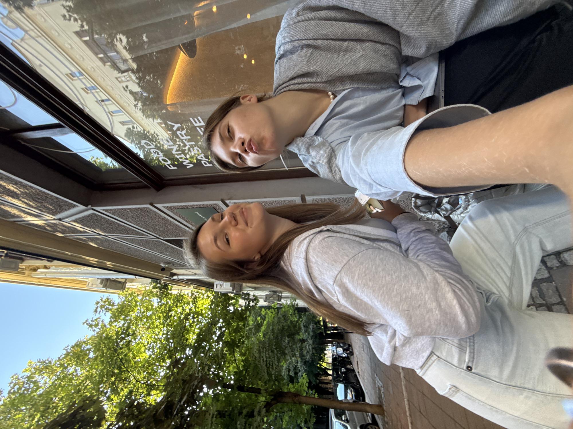 Two young women are posing for a selfie on a sidewalk in front of a building in Belgrade, Serbia. The woman on the left is wearing a light gray hooded sweatshirt and light blue jeans, and she is smiling at the camera. The woman on the right is wearing a light blue button-down shirt and a gray scarf, and she is making a kissing face at the camera. Behind them, the building has a large window with text that appears to read "FOOD WINE PASTRY CAFE". Trees line the sidewalk, and parked cars are visible in the background. The sun is shining, casting shadows on the sidewalk.