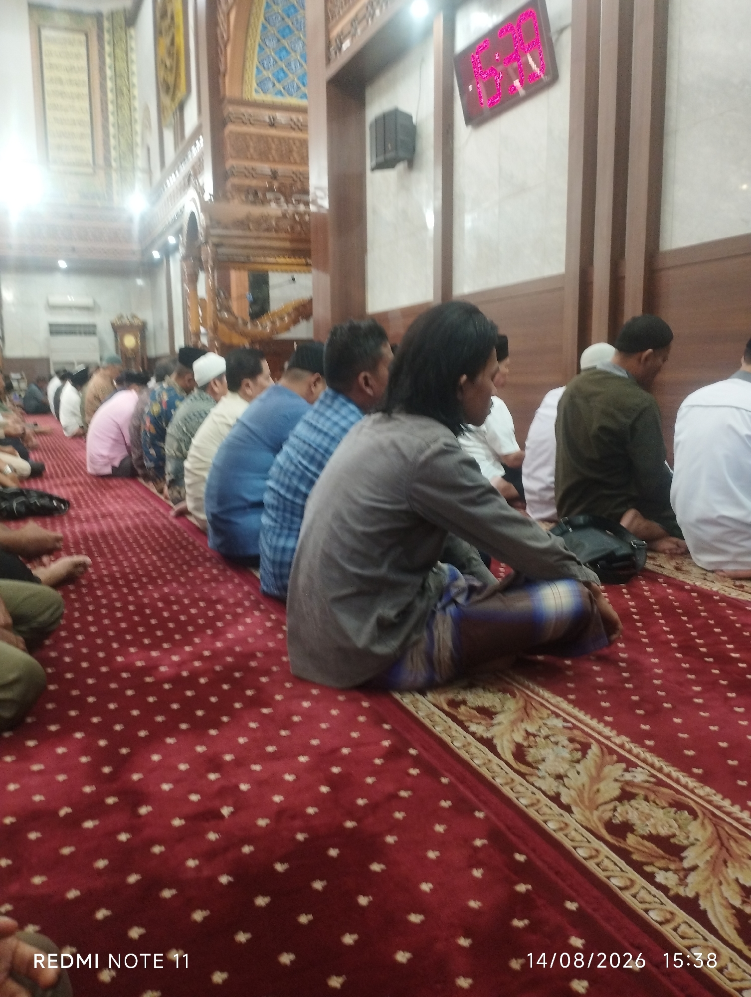 A group of men are seated on a red carpet with a patterned design, facing to the right of the frame, in what appears to be a mosque. The men are dressed in traditional Muslim attire. The room features ornate wooden decorations and large windows with decorative panes. A digital clock displaying "15:39" is visible on the wall. The timestamp on the image reads "14/08/2026 15:38", indicating the photograph was taken on August 14, 2026, at 3:38 PM. The text "REDMI NOTE 11" in the lower left corner suggests the image was captured using a Redmi Note 11 phone. The overall scene depicts a moment of prayer or reflection within a religious setting in Karawang, Indonesia.