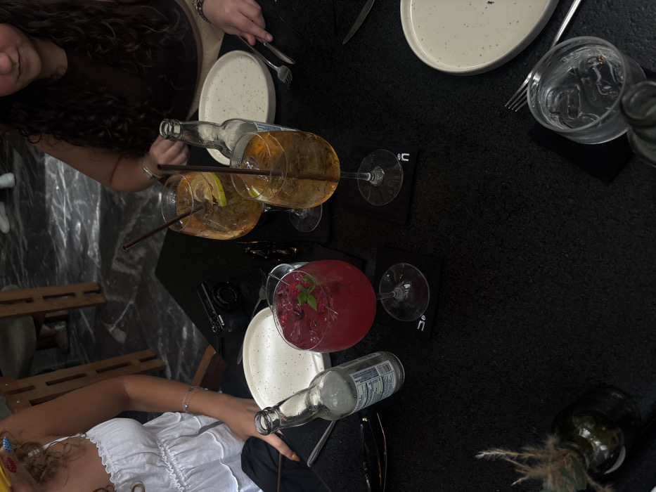 The image depicts an overhead view of a black, textured dining table occupied by two individuals in an indoor establishment in Unknown, Unknown. On the left, a person with curly hair, wearing a white ruffled top and a silver bracelet, has their right hand, with orange-brown painted fingernails, resting on the table next to a partially full clear glass bottle and a red cocktail garnished with raspberries and mint. On the right, a second person with long, dark curly hair, wearing a dark brown top, holds a fork and knife above an empty speckled ceramic plate. Two stemmed glasses, each containing an amber beverage with a lime slice and a straw, are positioned between the individuals. A black compact Canon camera is centered on the table. A clear glass of ice water and another empty speckled ceramic plate are visible on the bottom right. A pair of black sunglasses lies on the left, and a small dark green bottle containing dried wispy plant material serves as a decoration on the bottom left. The background reveals a dark, textured floor and wooden furniture.