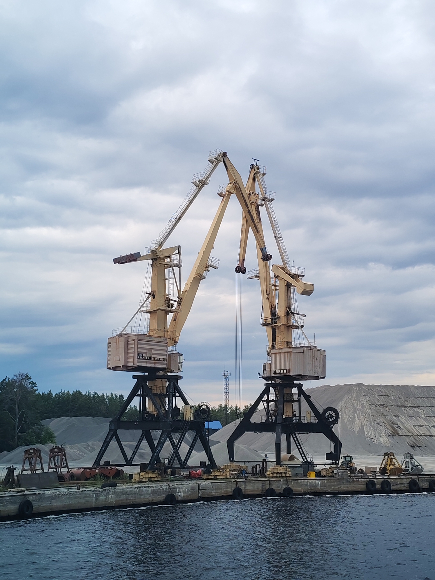 The image depicts a port area, likely in Shalsky, Russia, featuring two large, industrial yellow cranes positioned side-by-side on a concrete pier. The cranes have robust black metal supporting structures and long, angled booms. One crane has a visible label that appears to read "KRANBAU SEERSWAICE". The pier is laden with various objects, including what look like spare parts, barrels, and possibly equipment like a yellow bulldozer and a yellow excavator with a grapple attachment. In the background, large piles of gravel or sand are visible, along with a dense line of evergreen trees. The sky is overcast with dramatic, gray clouds. The water in the foreground is dark and rippling. There are no visible people in the image.