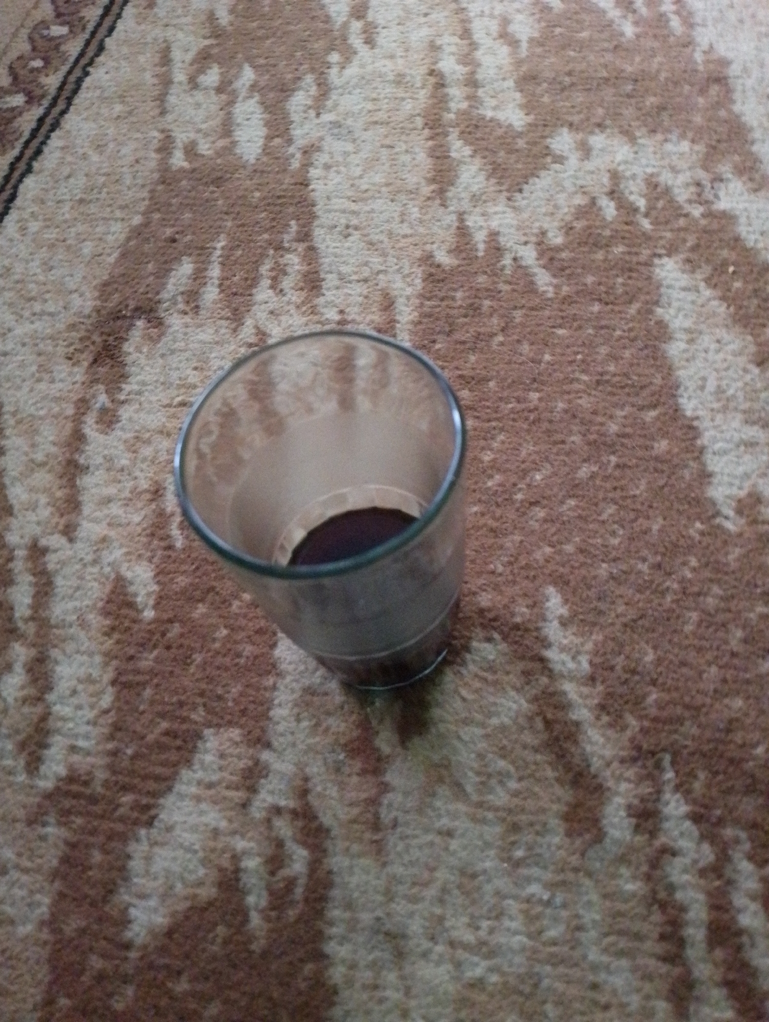 In Sukamanah, Indonesia, a clear glass tumbler containing a dark liquid rests on a carpet with a patterned design of brown and cream. The tumbler is positioned in the lower-left quadrant of the frame, at an angle. The carpet's pattern is abstract, with irregular shapes and streaks. The lighting suggests an indoor environment.