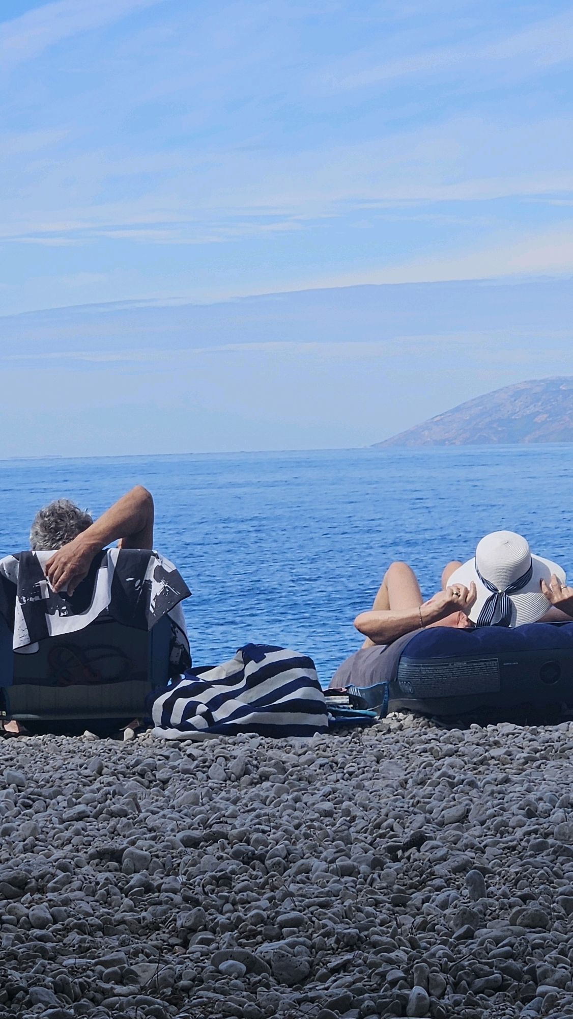Two individuals are relaxing on a pebbled beach in Općina Podgora, Croatia, with the Adriatic Sea stretching out before them under a clear blue sky. On the left, a person with gray hair sits in a chair, facing away from the camera, with their left arm resting on the chair's back. A black and white patterned towel is draped over the chair. To their right, a person wearing a white wide-brimmed hat with a navy and white striped ribbon lies on their back on an inflatable mattress, facing towards the sea. Their arms are resting behind their head and near their face. A blue and white striped towel is laid out in front of the inflatable mattress. The beach is covered in smooth, grey pebbles, and the water is a vibrant blue. In the distance, a hazy, mountainous coastline is visible.