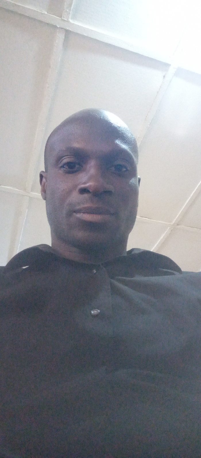 The image features a close-up portrait of a man, taken from a low angle looking upwards. The man has a dark complexion, a bald or closely shaved head, and appears to be in his adult years. He has dark eyes and a neutral or slightly serious expression, looking directly at the camera. His lips are closed.
He is wearing a dark-colored, possibly black or very dark grey, collared shirt or tunic. Two metallic-looking buttons are visible on the placket of his shirt, one higher up near the collar and another below it. The fabric of his shirt has a slightly textured appearance.
The background is a brightly lit, light-colored surface, likely white or off-white, with visible structural lines or beams forming a grid-like or panelled pattern. This suggests an indoor setting, possibly a ceiling or an upper wall. The lighting appears bright and somewhat diffused, illuminating the background significantly, and casting some soft highlights and shadows on the man's face. The overall composition places the man prominently in the foreground, filling the majority of the frame from just above his head down to his chest. No text is visible in the image.