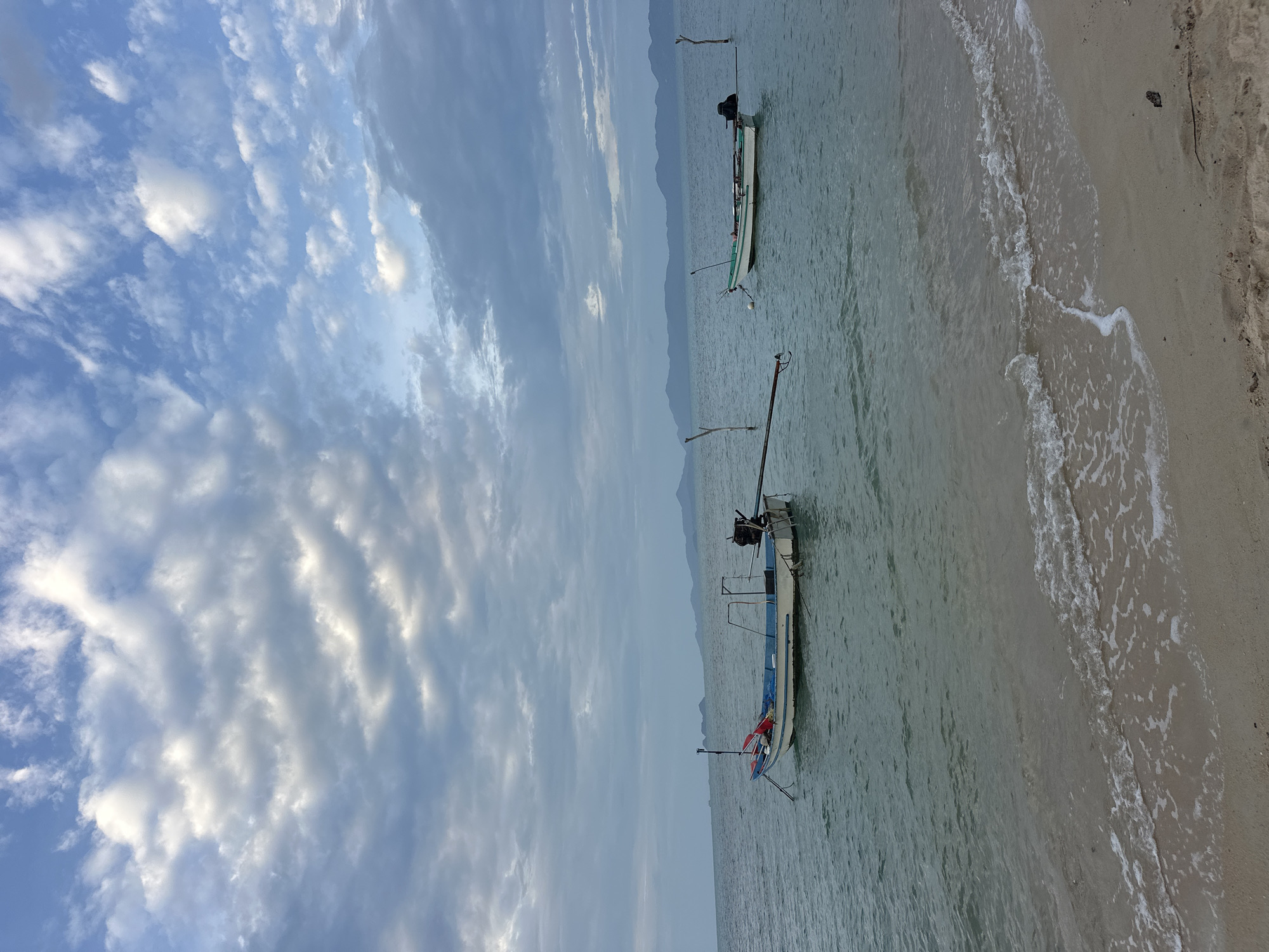 The image captures a serene coastal scene in Ko Samui District, Thailand. Two long-tail boats are anchored in the calm, turquoise waters of the Gulf of Thailand. The foreground features a sandy beach with gentle waves lapping at the shore. In the background, a hazy mountain range stretches across the horizon under a cloudy sky. The sky itself is a dramatic mix of blue and grey clouds, with patches of sunlight breaking through. The overall atmosphere is tranquil and picturesque, characteristic of a tropical beach setting.