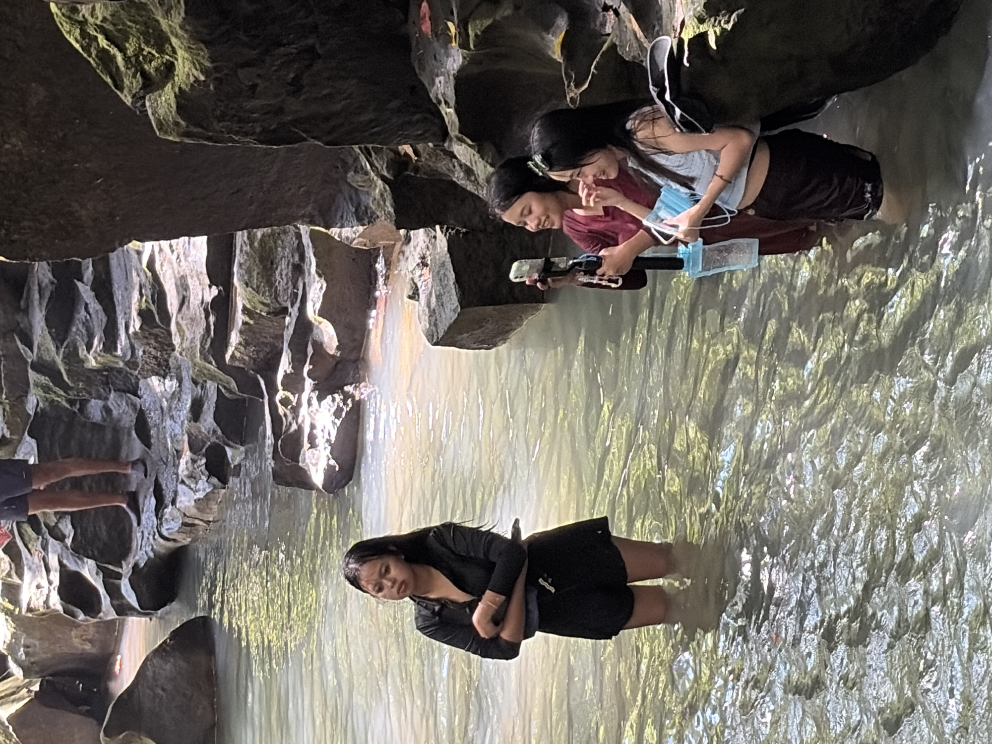 In Gianyar, Indonesia, at a jungle waterfall, three young women stand in shallow, clear water. The woman on the left, dressed in a black long-sleeved shirt and black shorts, stands with her arms crossed, looking to her left. To her right, two women are taking photos. The woman in the middle wears a maroon top and holds a selfie stick with a phone. The woman on the right, in a light blue sleeveless top and black shorts, smiles while holding a blue face mask and a water bottle. The environment consists of layered rock formations and lush greenery, with sunlight filtering through the canopy, creating dappled light on the water's surface.
