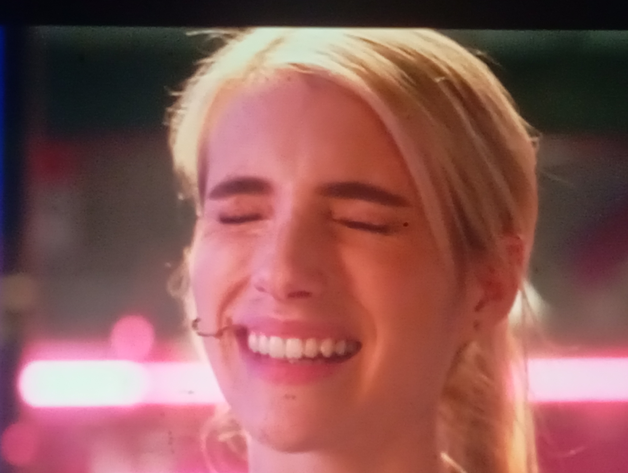 The media file is a close-up, low-resolution still image of a young Caucasian woman with blonde hair. She is smiling with her eyes closed, her teeth visible. She appears to be outdoors, with blurred neon lights in the background, suggesting a nightlife or entertainment setting. The lighting is predominantly pink and purple, casting a glow on her face. There are no visible objects or text. The woman's expression conveys happiness or amusement. The time of day is likely evening or night due to the artificial lighting and blurred background. There is no information about the weather. The location context provided is Sofiivska Borshchahivka, Ukraine, but there are no visual cues within the image to confirm this location.