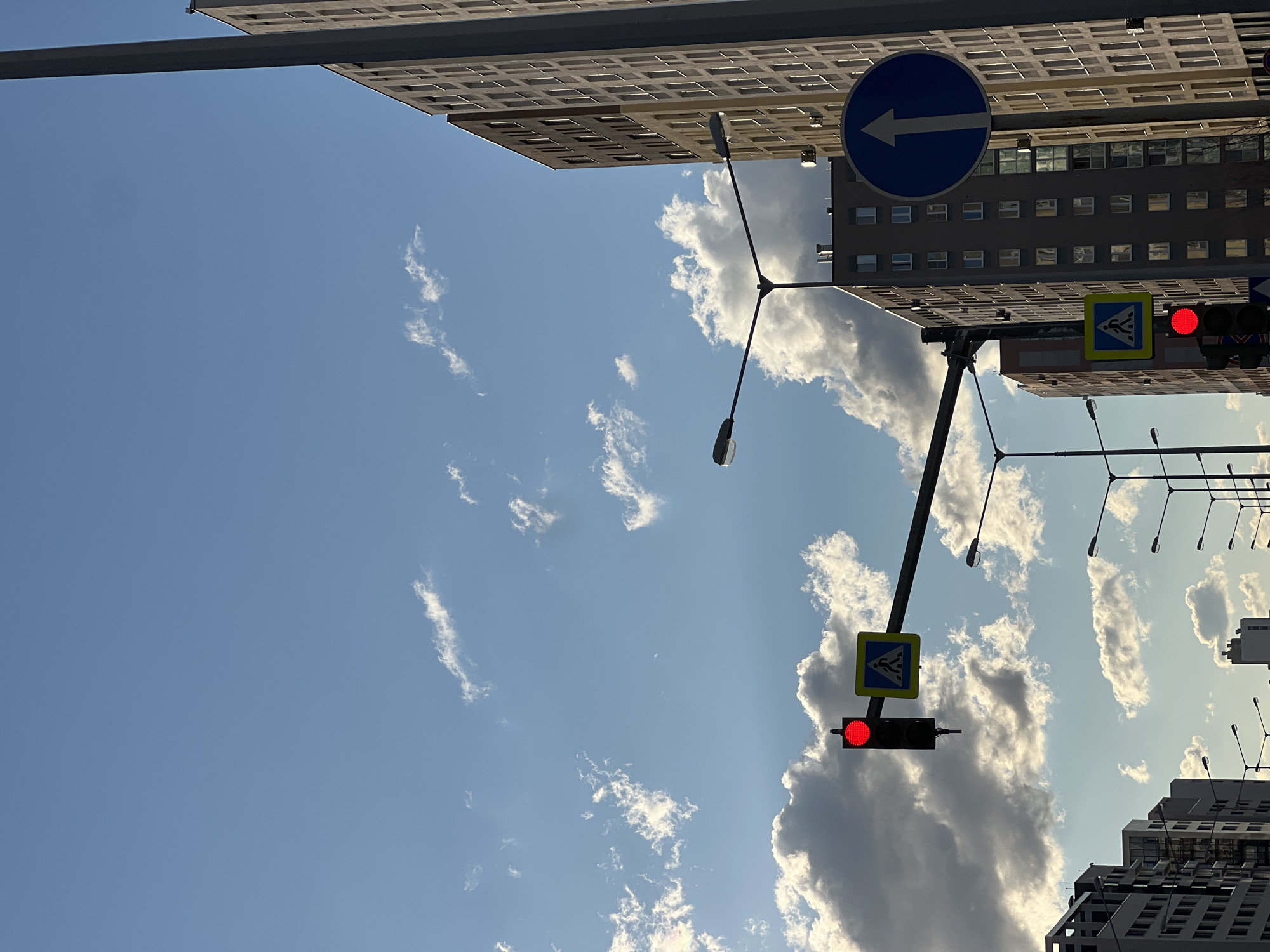 This media file captures a street scene in Yekaterinburg, Russia, under a bright, partly cloudy sky, suggesting it's daytime. The perspective is looking upwards, emphasizing the architecture and the sky.
The primary subjects are buildings, a traffic light, street lamps, and road signs. Two large buildings dominate the upper part of the frame, characterized by their numerous windows and grid-like facades. A traffic light is prominently displayed, showing a red signal, indicating vehicles should stop. Adjacent to the traffic light is a pedestrian crossing sign, a yellow square with a blue border and a white silhouette of a person walking. Another circular blue road sign with a white arrow pointing left is visible, indicating a mandatory direction. Several street lamps with a modern design are also present, extending from black poles.
The scene appears to be at an intersection or a busy street given the traffic signals and signs. The sky is a vibrant blue, dotted with scattered white clouds, some of which are dense and dramatic, while others are wispy. The lighting suggests a sunny day, casting shadows and highlighting the textures of the buildings. No people are visible in the frame. The overall mood is urban and functional, with a focus on the infrastructure of the city. No text is discernible on the buildings, signs, or traffic lights, other than the standard symbols on the road signs.