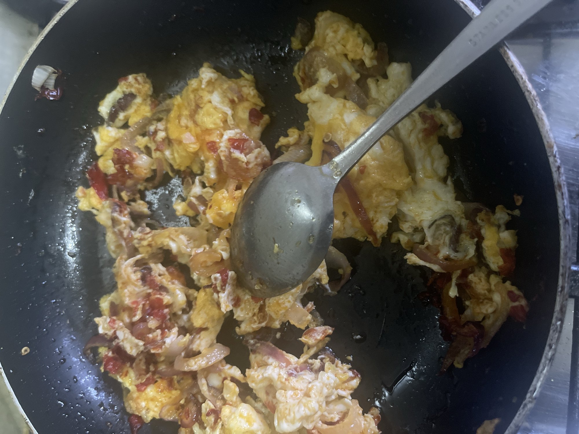 A top-down, close-up shot shows scrambled eggs with red onions and red peppers being cooked in a non-stick black frying pan on a stovetop. A stainless steel spoon rests in the pan amongst the eggs. The environment around the stove appears to be a kitchen setting. There is no indication of the specific location within Rafin Bauna, Nigeria. No people are visible.