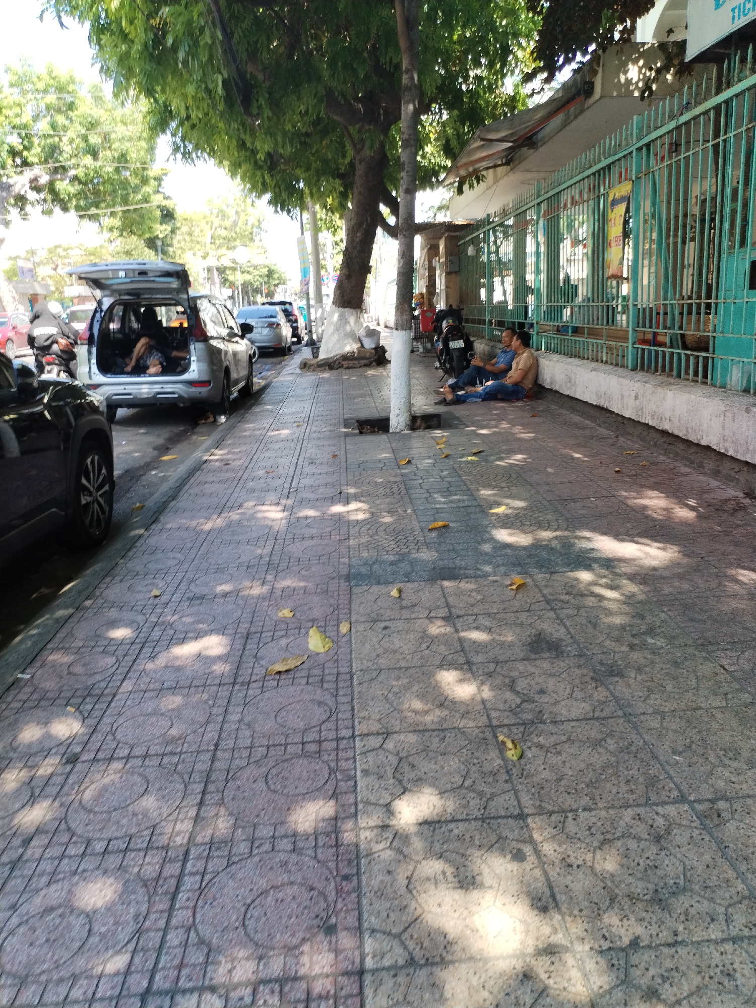 The image depicts a sidewalk in Nha Trang Ward, Vietnam, with a row of parked cars and motorcycles along the street. A silver minivan has its rear hatch open, revealing two individuals inside, one lying down. Two men are seated on the sidewalk, leaning against a green metal fence. The sidewalk is paved with patterned tiles and has fallen leaves scattered across it. Shadows from trees fall on the ground. A tree trunk is painted white at its base. A gutter runs along the edge of the sidewalk.