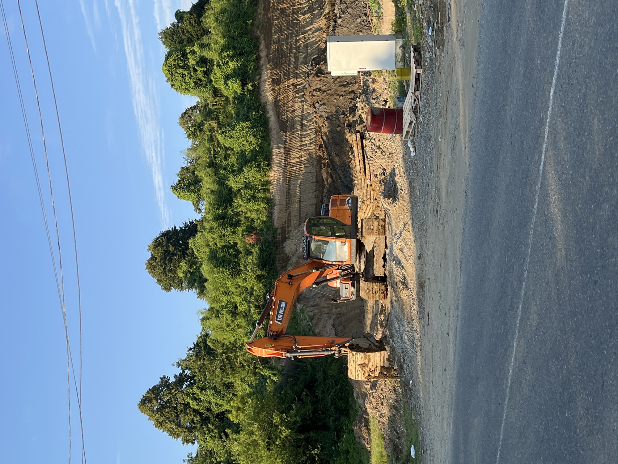 An orange excavator is parked on a dirt and gravel construction site. The excavator is facing right, with its arm lowered and bucket resting on the ground. Behind the excavator, a large embankment of earth has been excavated, revealing layered soil and rock. Trees and green foliage cover the top of the embankment. In the foreground, a paved road runs horizontally across the bottom of the image. A red barrel and a white utility box are visible on the right side of the construction site, near a wooden pallet. The sky above is clear and blue, with a few thin white clouds. The scene appears to be set in a rural or semi-rural area, possibly on the outskirts of Ureki, Georgia, given the presence of construction and natural vegetation.