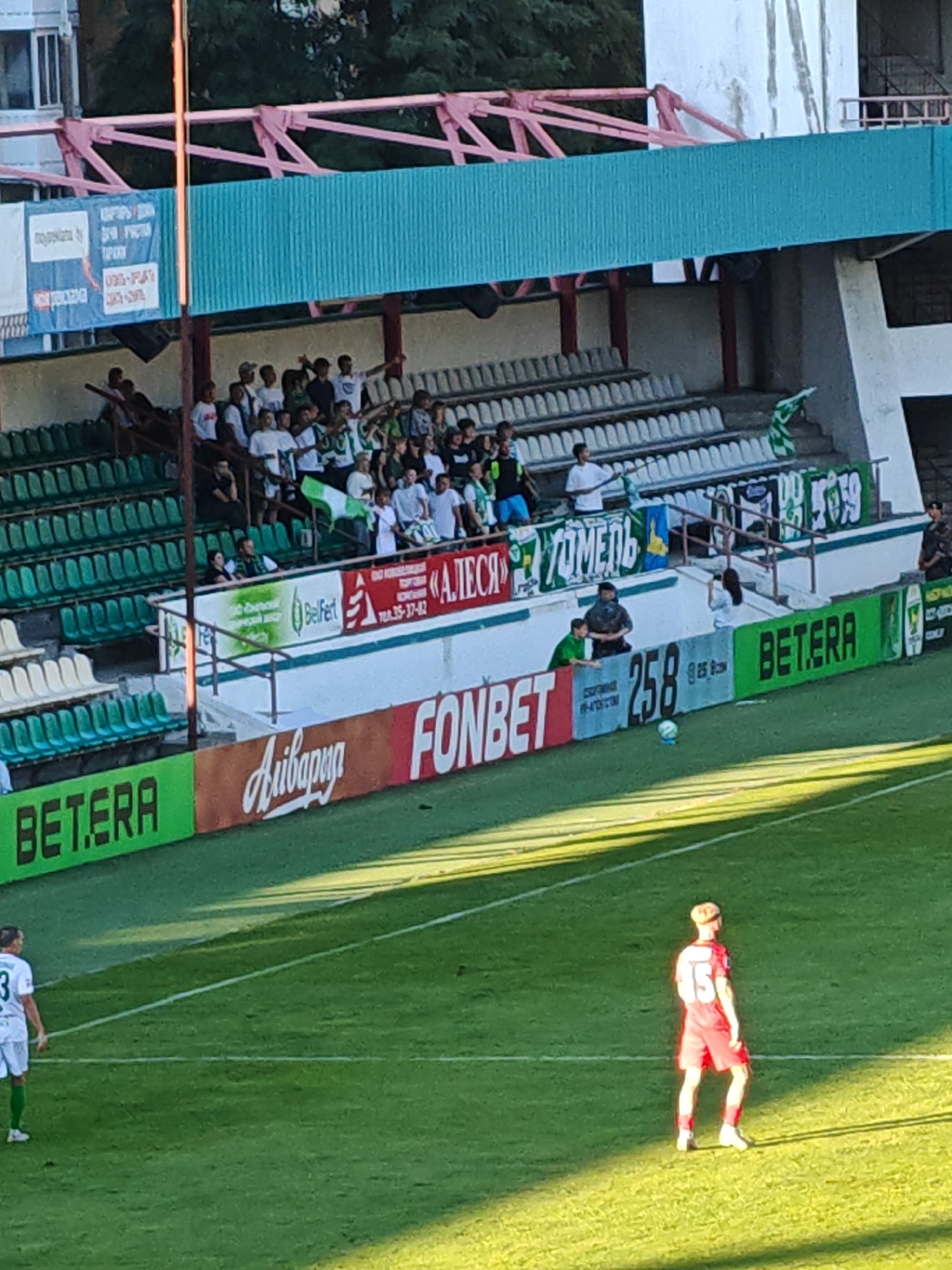 The image shows a view of a sports stadium with a football match in progress. Spectators are seated in the stands, and players are on the field. The stadium features green seating and a turquoise-colored roof structure. Several advertising banners are visible along the lower section of the stadium, including "BET.ERA" and "FONBET". A football is visible on the field, and a player in a red uniform with the number "15" on his back is standing on the grass. Another player in a white uniform is further to the left. The overall scene suggests a sports event taking place in an outdoor stadium, likely in Homyel, Belarus, given the context of the advertisements and stadium design often associated with the region.