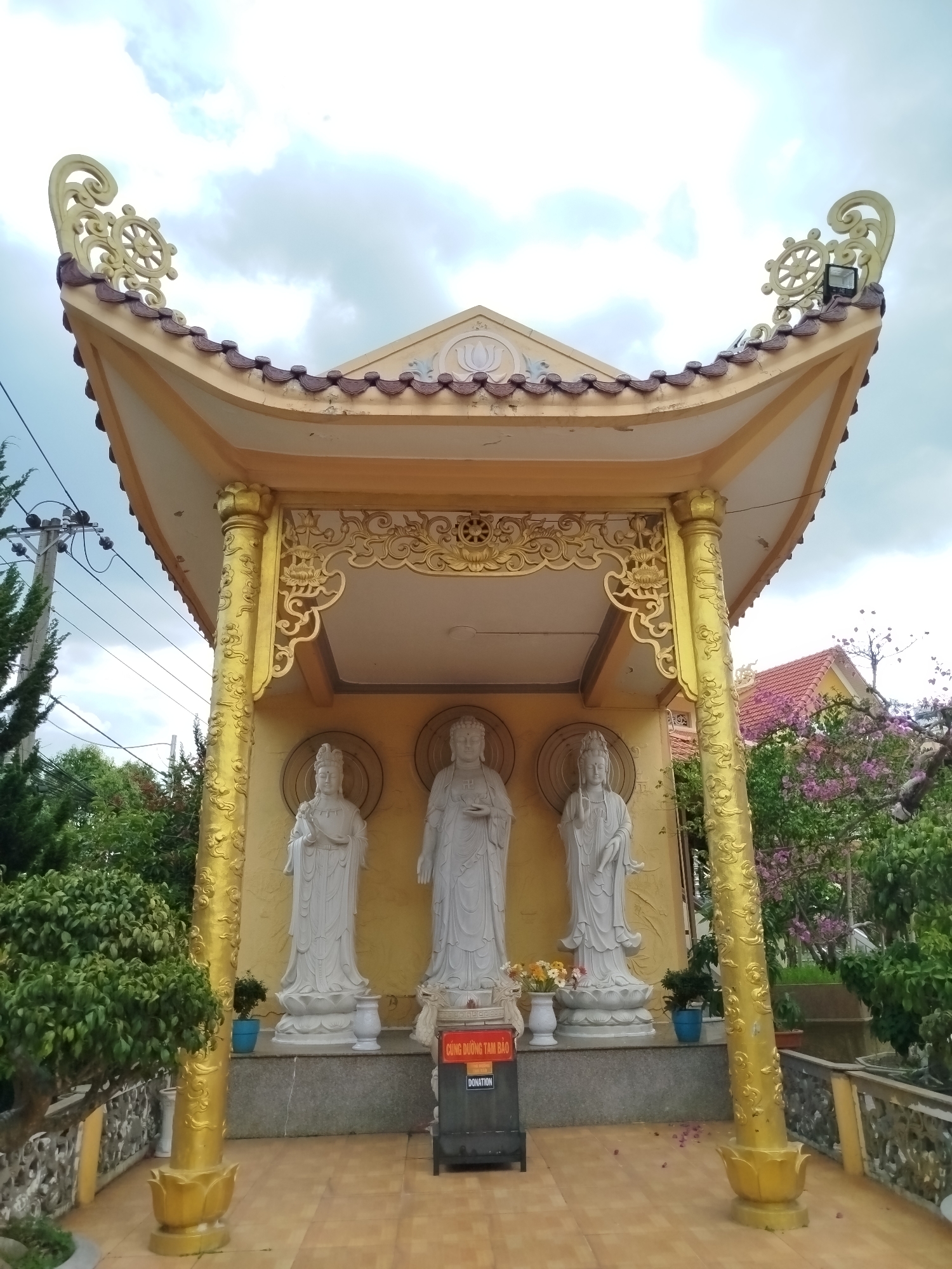 The image depicts a Buddhist shrine located in Lâm Viên, Đà Lạt, Vietnam. The shrine is a small, ornate structure with a curved, tiled roof and gilded decorations. Within the shrine are three white statues of deities, each standing on a lotus pedestal. A donation box is placed in front of the statues. The shrine is surrounded by greenery, including trees and flowering plants. The sky is overcast with scattered clouds.