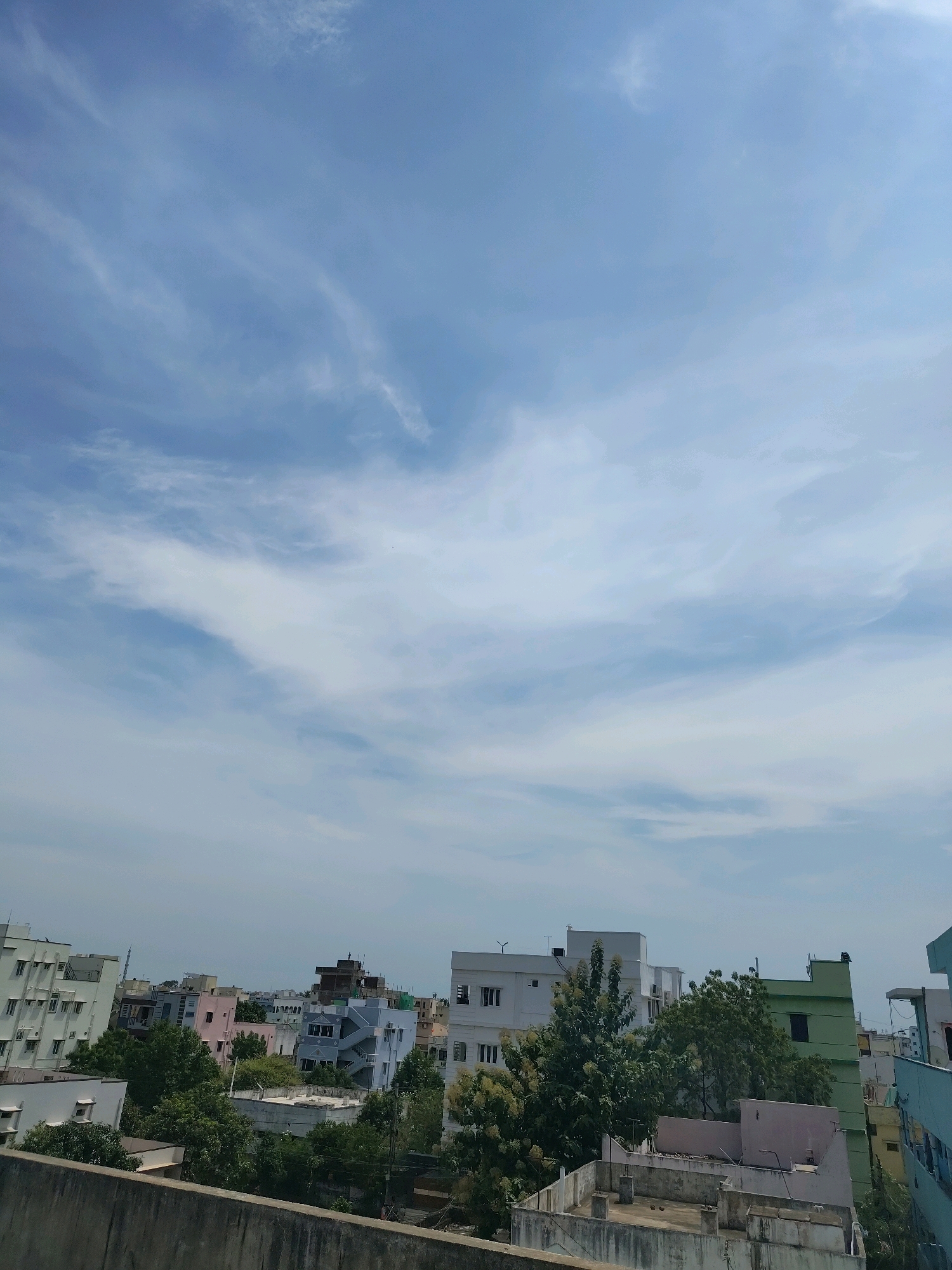 This is a ground-level, outdoor, medium-shot photograph captured on a bright, sunny day, depicting a cityscape. The image is oriented vertically and is dominated by a clear blue sky with scattered, wispy white clouds. In the foreground, a concrete wall runs horizontally across the bottom of the frame, providing a sense of perspective. Beyond the wall, a collection of buildings of various heights and colors can be seen, some appearing to be residential and others commercial. These structures are interspersed with lush green trees, creating a contrast between the built environment and nature. The architecture suggests a South Asian setting, possibly in the city of Satyanarayana Puram, India, given the characteristic building styles and the overall atmosphere. There are no visible people in the frame, and the scene appears to be a typical urban landscape. The lighting is bright and even, indicating midday.