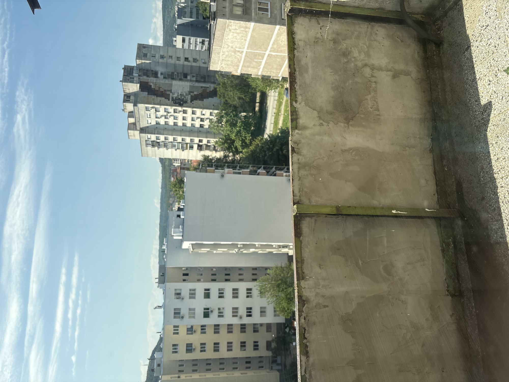 The image, taken from a high vantage point in Lovech, Bulgaria, shows a view of the city’s residential area. Concrete apartment buildings dominate the scene. The sky is clear with some clouds. A concrete balcony edge with a corroded metal beam is visible in the foreground.