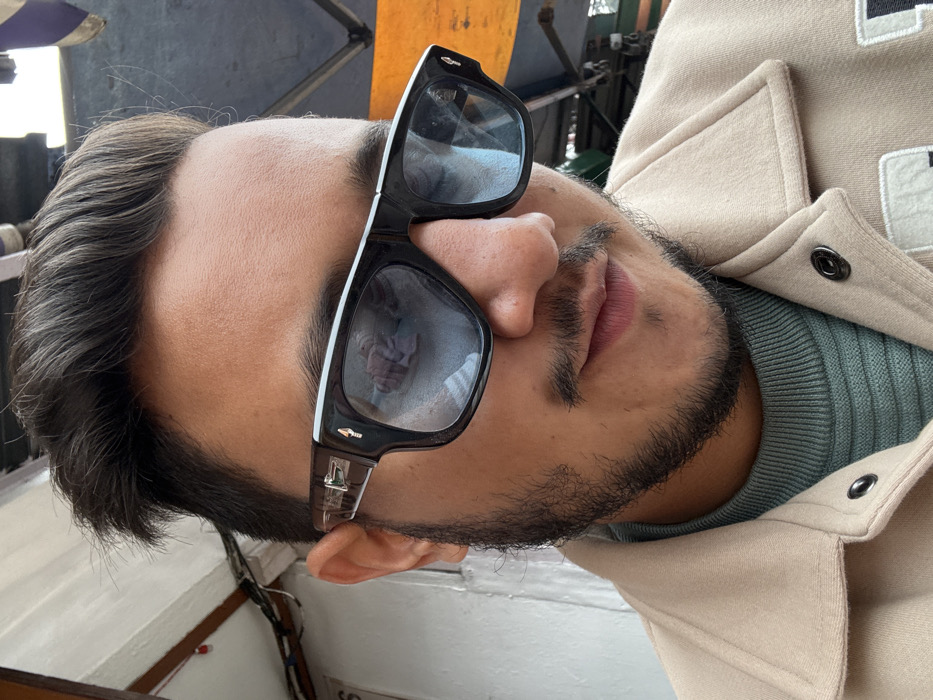 A male individual is featured in a close-up portrait, positioned outdoors in Mussoorie, India. He has dark brown hair styled with volume, and a short beard and mustache. He wears black-framed sunglasses with white accents along the upper edge and metallic details on the temples; the lenses are light blue tinted. Reflections in the sunglasses show a blurred human figure and indistinct background elements. The subject is dressed in a ribbed teal turtleneck sweater, layered under a light beige or tan collared jacket secured with visible dark snap buttons. His head is slightly tilted, and his gaze is directed downwards and to the right. The blurred background indicates an urban street setting, with a light-colored wall or structure partially visible on the left, and elements suggestive of an overhead roof structure or awning with wiring, alongside segments of an orange/brown tarp or canopy in the upper right.