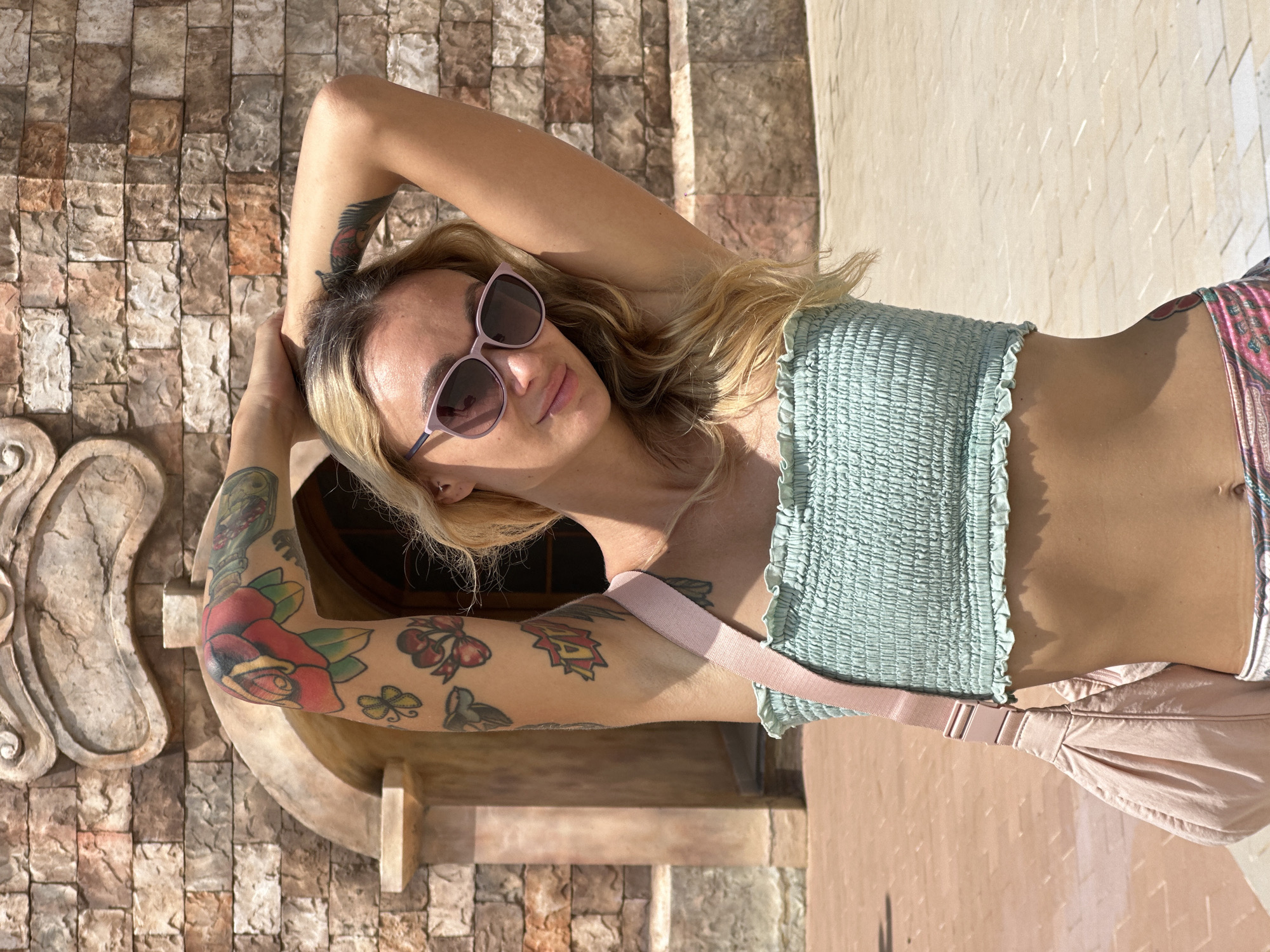An outdoor, sunlit, full-length portrait in Tien Thanh Ward, Vietnam, features a light-skinned woman positioned centrally, looking towards the viewer with a slight smile and her head tilted to her left. Her long, wavy, blonde hair, with visible darker roots, cascades over her shoulders. She wears pink-framed sunglasses with dark lenses, a light green smocked strapless crop top that exposes her midriff, and patterned lower garments in shades of pink, green, and white. Her arms are raised, with her hands clasped behind her head, revealing a variety of colorful tattoos: a large red rose with green leaves on her right elbow, a yellow four-leaf clover and red cherries with a bow on her right forearm, and a feather-like design on her left bicep. A light pink, soft-sided cross-body bag with a wide strap and plastic buckle hangs over her right shoulder. The background consists of a building facade constructed from multi-toned rectangular stone blocks, featuring a central archway and an ornate, sculpted stone relief above it. The ground is paved with light-colored rectangular pavers, and strong shadows indicate bright, direct sunlight.