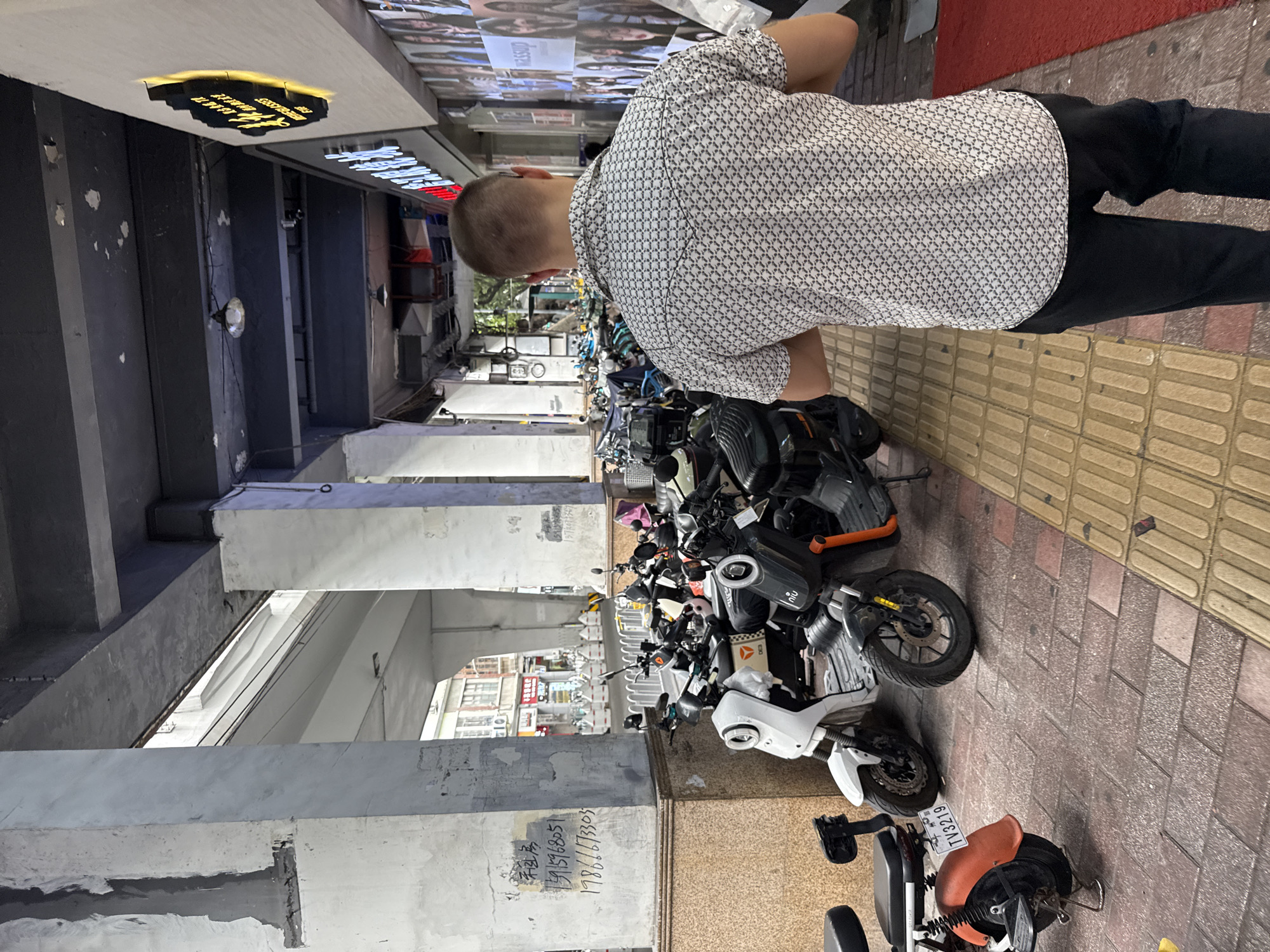 In Yuexiu District, China, a man with a short buzz cut is walking away from the camera. He is wearing a white short-sleeved shirt with a repeating black geometric pattern and black pants. The man is walking on a sidewalk paved with red and yellow textured tiles. To his left, a row of electric scooters and motorcycles are parked. Some of these vehicles are white, black, and orange. One of the white scooters has a license plate that reads "TV3219". To his right, a building features a wall covered with posters. Above the sidewalk, a concrete overpass structure is visible, with thick concrete pillars supporting it. The environment appears to be an urban area, possibly a commercial street, with buildings and infrastructure characteristic of a city. Graffiti is present on one of the concrete pillars.