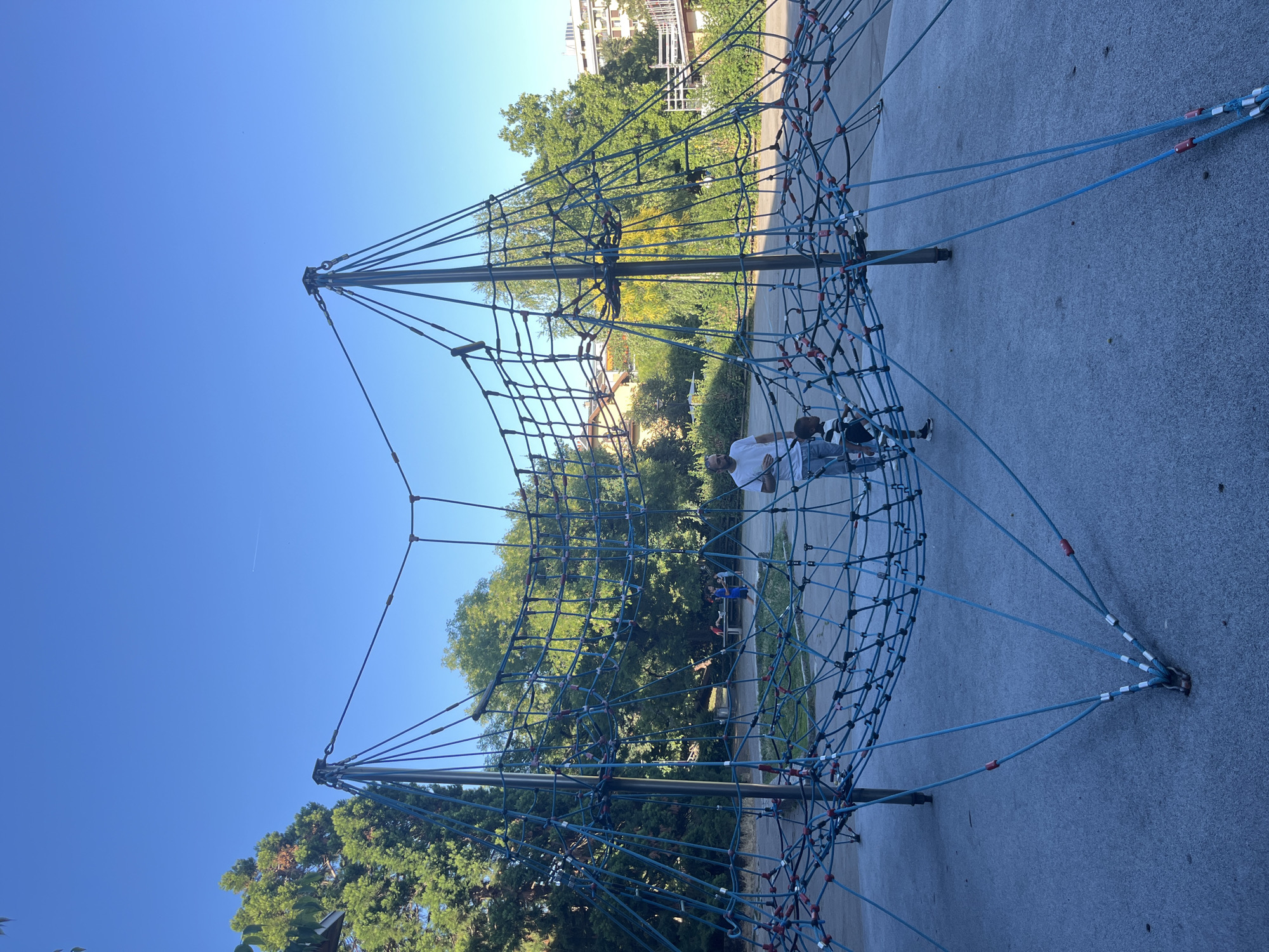 The image shows a large, blue rope climbing net structure in a park. A man in a white t-shirt and a child in a striped shirt are standing on the net. The park has trees, a blue sky, and buildings in the background. The ground is paved with a grey material. The scene appears to be in Geneva, Switzerland, based on the architecture of the buildings and the style of the park.