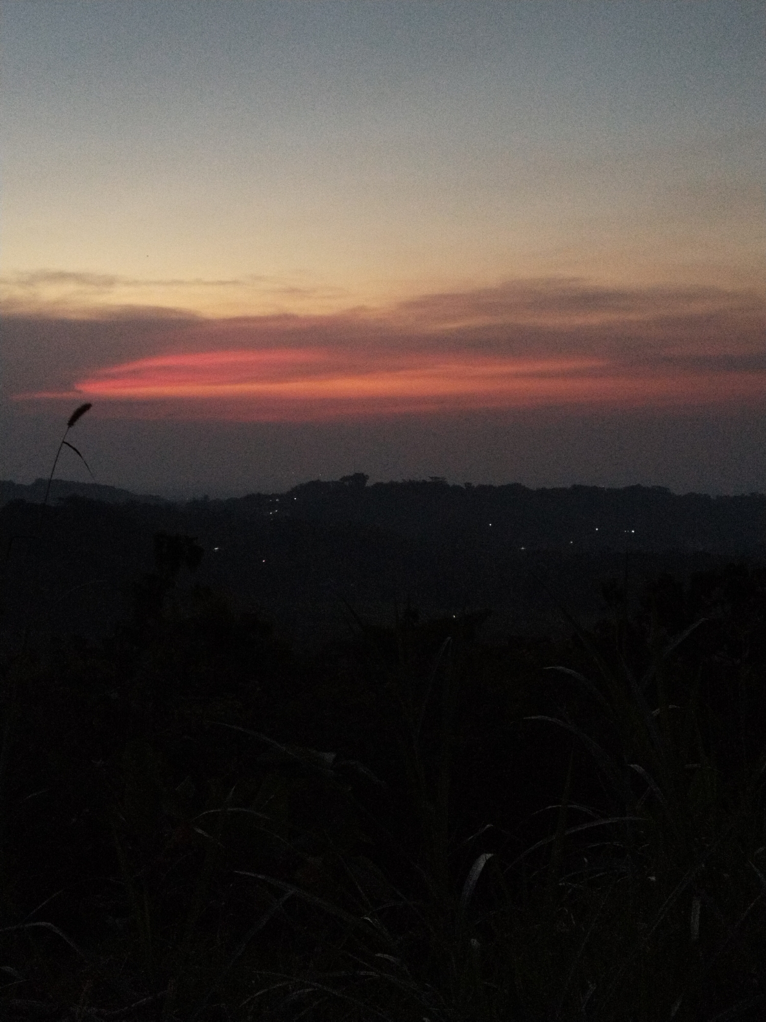 The image depicts a dusk or dawn scene with the sky transitioning from a dark blue at the top to a peach hue, and finally to a band of red near the horizon. Below the horizon, a dark silhouette of a treeline or hillscape is visible, dotted with faint lights, possibly from buildings or streetlights. In the foreground, there are dark, indistinct shapes suggesting tall grass or vegetation. The location is implied to be a city named Unknown within the region of Unknown.