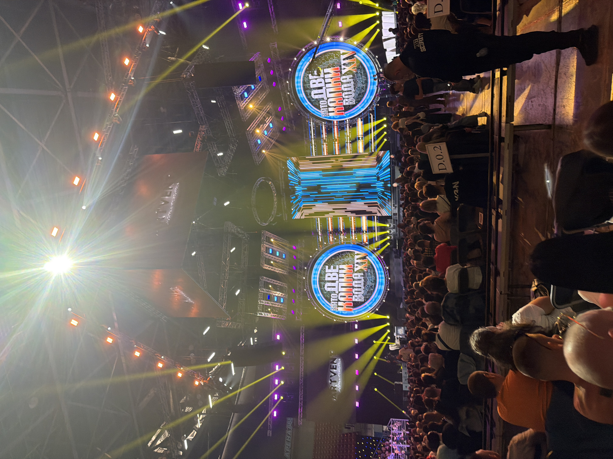 Here is a detailed description of the image:
**Scene:** The image captures a view from a concert or event held in Arena Sofia, Bulgaria. The arena appears crowded with audience members, who are mostly facing a large stage.
**Stage and Lighting:** The stage is elaborately decorated with lighting structures, including yellow, purple, and white beams. There are two circular signs with blue and white lettering on the stage. One sign says "ДВЕ КАПКИ ВОДА XIV" (Two Drops of Water XIV) and another sign appears to have text on it.
**People:** A large crowd fills the seating area, with visible heads and shoulders of many individuals. Nearer to the photographer, there are several audience members. A man with "WEST SECURITY" on his black shirt is standing by the crowd.
**Objects:** Besides the stage structures, lighting, and signage, there is a hand-held sign with the text "D.0.2" printed on it.
**Environment:** The scene is indoors and has the distinct atmosphere of a live performance.