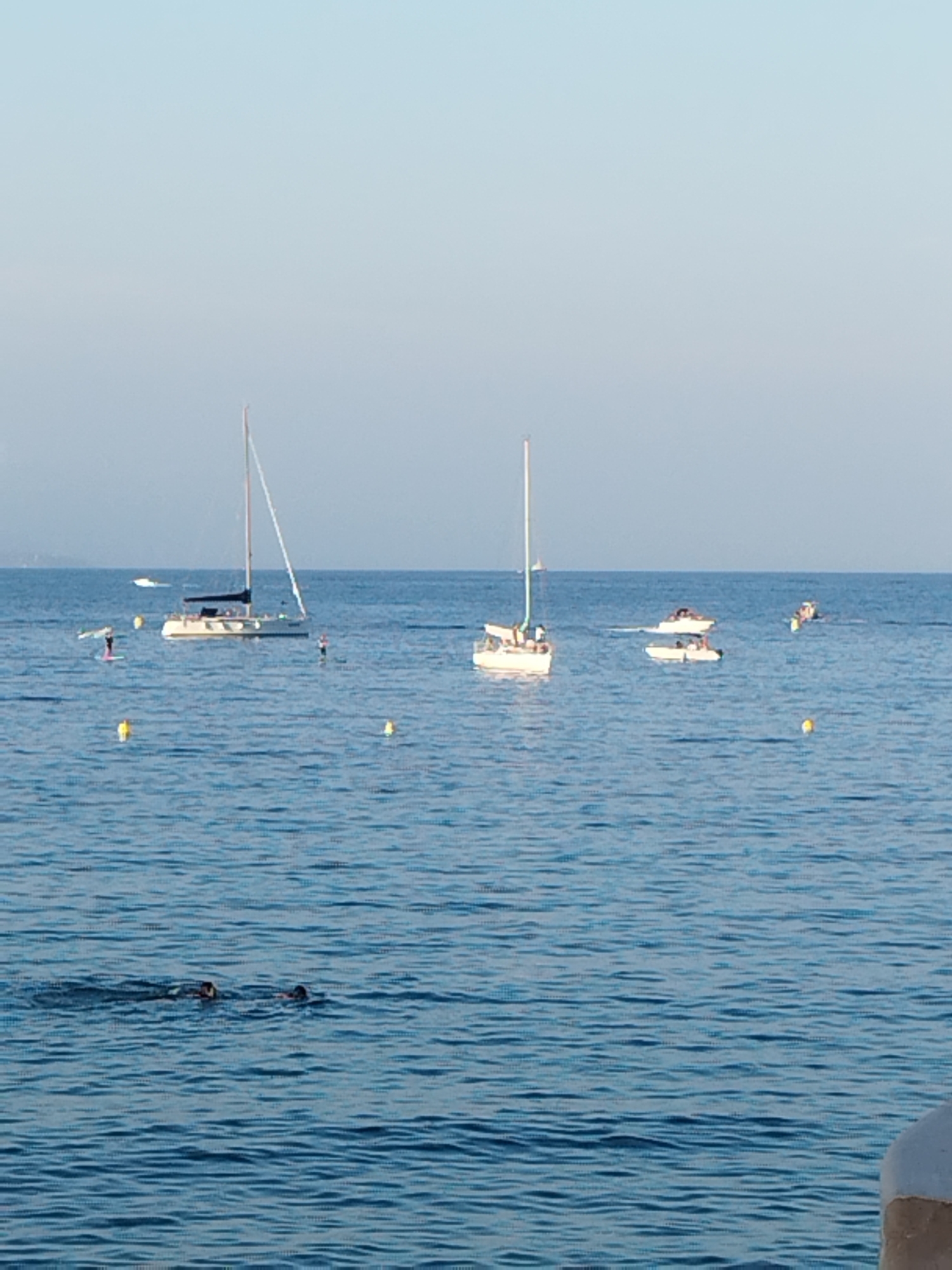 A wide-angle, eye-level, long shot captures a clear day at sea in Antibes, France, with a tranquil blue ocean under a pale blue sky. In the foreground, two people are swimming, their heads and shoulders visible above the water. In the mid-ground, several boats are scattered across the water. A prominent sailboat with a white hull and mast is positioned to the left of center. To its right, two smaller motorboats are visible, one carrying passengers. Further in the distance, other boats and buoys are present. The scene suggests a leisurely day spent enjoying the Mediterranean Sea, with both recreational swimming and boating activities taking place.