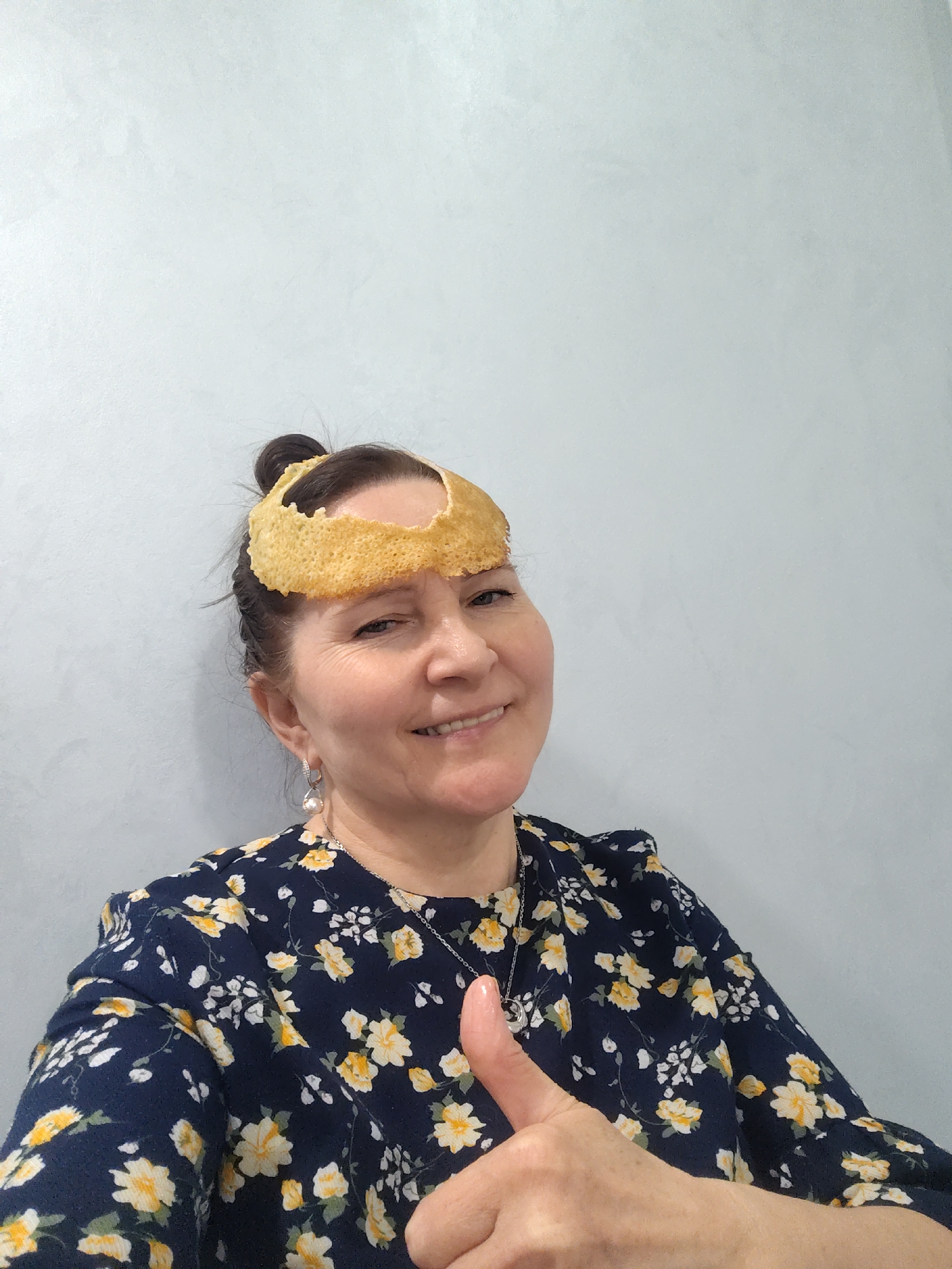 A woman with brown hair styled in a bun is smiling and giving a thumbs up. She is wearing a dark blue floral dress with yellow and white flowers and a silver necklace with a pendant. She also has a pair of dangling pearl earrings. On her head, she is wearing what appears to be a crispy, circular food item, possibly a cracker or a thin, fried cake. She is standing against a light grey or white wall.
There is no information to determine if this image was taken in the city of Sarapul, Russia.