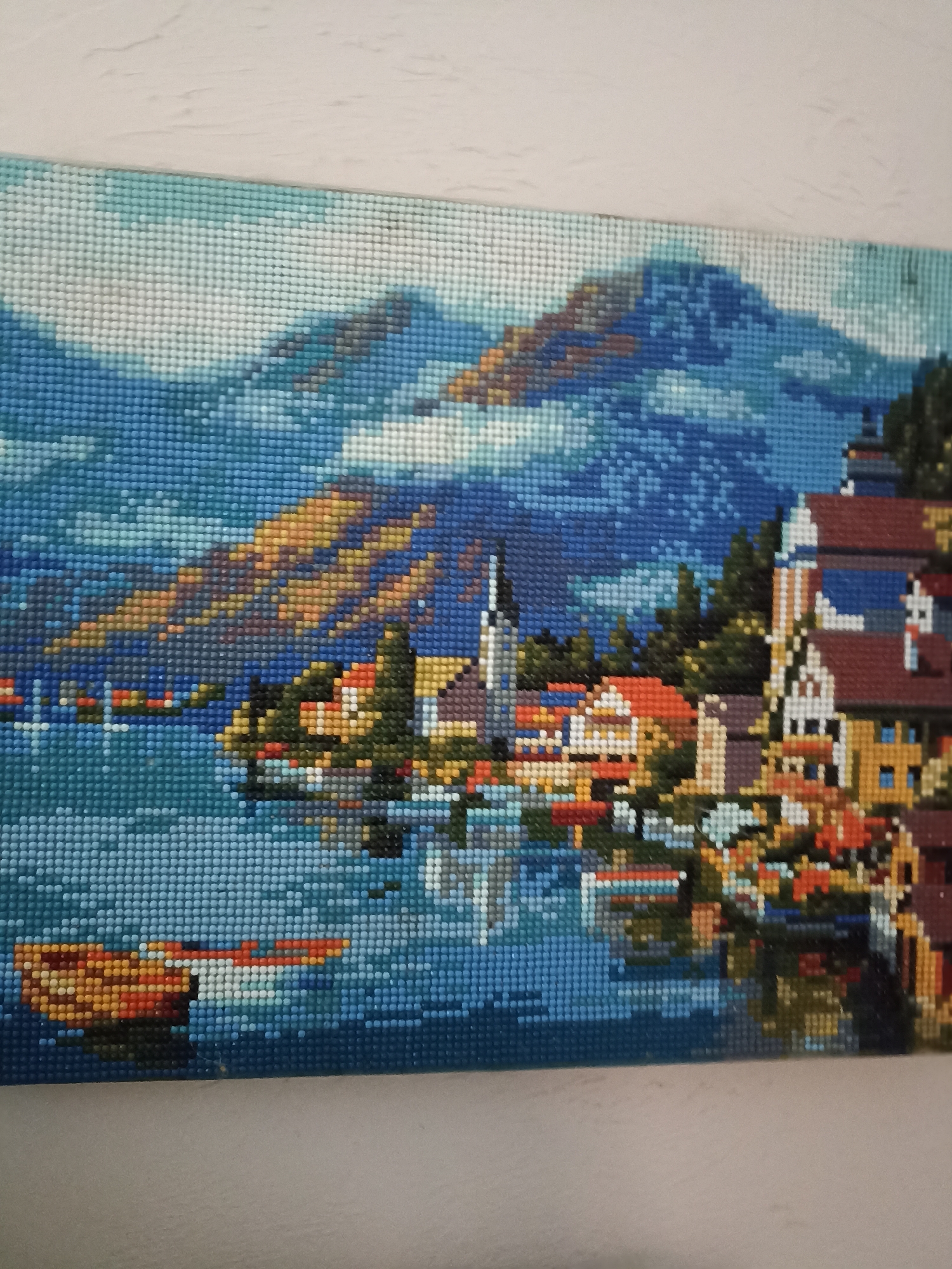 The image displays a diamond painting of a panoramic landscape. The painting depicts a village situated on the bank of a large body of water, likely a lake, at the base of prominent mountains. The visual characteristics of the depicted village, including its architecture and mountainous lake setting, are consistent with the well-known Austrian village of Hallstatt, not Velykodolynske, Ukraine.
In the foreground, a calm expanse of blue water occupies the lower half of the painting, featuring pixelated reflections of the village and mountains. A distinct orange and brown boat is positioned in the lower-left section of the water.
The midground features a cluster of buildings characteristic of European alpine architecture, including structures with red-tiled roofs, white or cream facades, and various brown and yellow details. A prominent church, distinguished by a dark, pointed spire, is identifiable within the village towards the left side of the cluster. Green trees are interspersed among the buildings and extend up the lower slopes.
The background is dominated by large mountains rendered in shades of blue, brown, and hints of orange, suggesting rugged terrain. Portions of the mountains and sky contain lighter blue and white pixels, indicative of clouds or mist. The sky above the mountains is predominantly blue. The entire artwork is composed of numerous small, uniformly sized square or round resin beads that form a mosaic-like image.