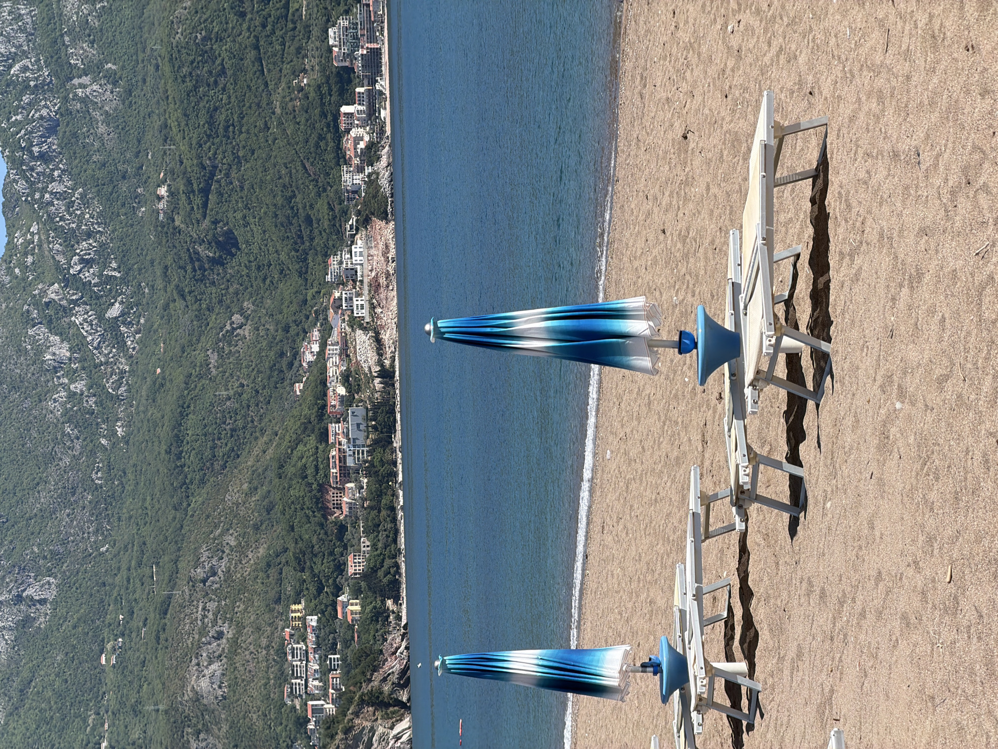 The image depicts a beach in Boreti, Montenegro. The scene includes a coastline with a mountain range on the left and buildings along the water's edge. The beach itself is sandy, with beach chairs and closed blue-and-white umbrellas. The sea is a deep blue color. There are no people visible in the image. The lighting suggests it is daytime.