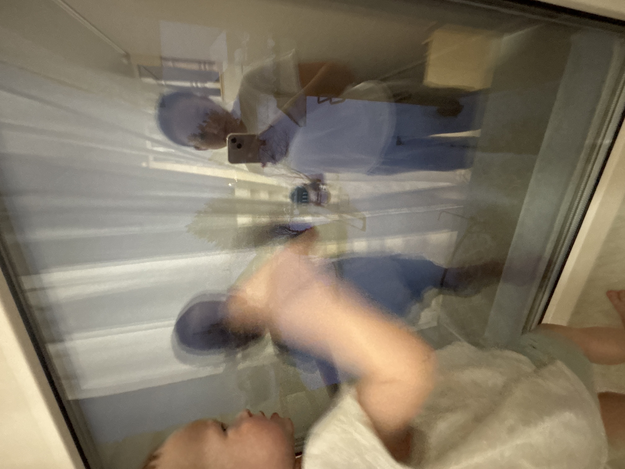 The image depicts a reflection in a window or glass door. The reflection shows a person holding a smartphone, presumably taking the picture. They are wearing a light-colored top and jeans. The background of the reflection includes stairs. The foreground shows a person, possibly a child, with their face visible at the bottom, wearing a light-colored garment. Part of their leg is also visible. There is no information to identify this image in Ferma, Russia.